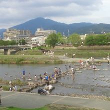 大勢の人が飛び石ジャンプで川を渡っています。後ろの山が比叡山