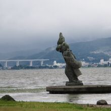 諏訪湖に浮かぶ像で、対岸の中央道岡谷高架橋が背景になっていま