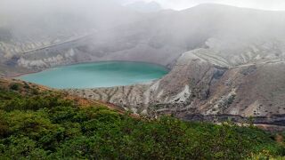 霧が一瞬晴れ、幻想的なエメラルドグリーンの火口湖が現れる。