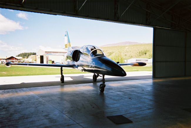 事業用操縦士のライセンスは持っているものの４人乗り小型プロペラ機セスナ社のC172しか操縦したことのない僕は、一度でいいからジェット機を操縦したいといつも夢見ていました。<br />そして2006年10月、ニュージーランド旅行をすることになった時に、現地での飛行機の体験操縦が出来る所を探していたところワナカでジェット戦闘練習機に搭乗が出来ることが分かりました。<br />正直言うと戦闘機はあまり好きではないのですが、ジェット機の操縦が出来る機会はなかなかないと思い申し込みました。<br />この旅行記はその体験記です、全体の旅行記は「ニュージーランド南島一人旅」をご覧ください。<br /><br />＜2006年10月19日＞ワナカ空港12:30離陸　12:55着陸<br /><br />フライトの予約は１４4時からだったのですが様々ないきさつ（旅行記「ニュージーランド南島一人旅【2】」参照）があり、１２時にJETFLIGTHS WANAKA社のグランツ教官と合流、ワナカ空港西隅の格納庫に向かいました。<br />歩きながら今日の天気の確認・・・今日は風がかなり強いが滑走路と正対の風なので問題なし（着陸後、やや右側から41ノットの強風と分かった）、雲も朝は多かったがだんだん晴れてきたとのでフライトに問題はないとの事。<br />暫く歩き格納庫に到着、早速扉を開くと・・・そこに居ました！！今日の搭乗機チェコスロバキア製のジェット戦闘練習機AERO L39Cアルバトロス(機体識別記号ＺＫ−ＬＬＲ)！！<br />練習機なので、最高速度４９１ノット、制限加重は＋８Ｇ〜−４Ｇ、実用上昇限度が11,000ｍと現役の戦闘機に比べるとかなり穏やかな性能ですが、私が普段操縦しているセスナC172と比べると（比べるのも恥ずかしいけど・・・）速度でも５倍とさすがの性能、基本的には名の通り練習機ですが、国によっては軽戦闘用に配備もされているそうです。<br />実は戦闘機を間近で見るのは初めて、その用途には嫌悪感を感じますが、その流麗なフォルムは美しいとしか言いようがありません。<br />荷物を格納庫に置き、用意されていたつなぎ服に着替えると気分は既に戦闘機乗り・・・さっきまで”戦闘機は用途が気に入らないんだよなぁ〜”なんて思っていたのにやはりワクワクは押さえきれません。<br />ついでにもし嘔吐した時の為のエチケット袋も渡されつなぎの足ポケットに入れておきます、結構なＧが掛かるので気分が悪くなる人もいるそうだ。<br />グランツ教官は牽引トラクターで機体を格納庫から芝生に押し出します、太陽の下で見るL39Cも美しい！！まるで生き物ような雰囲気！！<br />機体に刻まれた切り込みを足場にして後席に乗り込み、しっかりハーネスを締めると、教官がインストラクションを始めます。<br />要点は以下の通り・・・<br />・基本的に操縦は任せるが（教官は僕が日本の事業用ライセンスを所持している事を承知している）、離着陸は教官が行うので、その間は機器に触らないで欲しい、特にブレーキには触れないように。<br />・上空で機が安定したら通常の操縦交代手順（操縦を渡す方が「You have control」とコール、受ける方が「I have control」とコールし操縦を交代する）で操縦の受け渡しを行う。<br />・もし緊急事態が起こった場合は脱出レバーを引いて座席ごと射出するように。<br />の３点・・・もちろん初めての機種、しかもジェット戦闘機の離着陸なんて出来る訳もないので素直に了解の意を示します。<br />次に前席と後席での通話チェック、教官の「How dou you read?」の問い掛けに「Loud and clear.How do you read me?」と返答、教官の「Loud and clear」の応答でチェックは完了。<br />いよいよエンジンに火が入ります、前触れもなくゴーッッとエンジンがスタートします、普段聞き慣れたレシプロエンジンの音とは全く違うジェットエンジンの発する連続音はとても新鮮。<br />各種チェックを終えるとタクシー開始、暫く芝生を走りエプロンの舗装路面に乗り入れます、今日の使用滑走路はRWY29。<br />管制用の無線はよく聞き取れなったけどどうやらワナカ空港には管制機関がないようで、一方送信をしながら滑走路に乗り入れます。<br />滑走路を東端まで一旦進み180度ターンして向きを西に変えます。<br />「ZK-LLR Now Take Off Runway 29」教官が一方送信し、いよいよ離陸です、後ろでエンジン音が高まり機はダッシュを始めた。<br />思っていた程でもないけど明らかにセスナ172とは違う（あたりまえ！)加速！！あっという間に離陸速度に達し教官が操縦桿を引きます、機は軽やかに上昇します・・・が、対地200ft（約60ｍ）で早くもロール旋回（横回転の宙返り）を実施！！この低高度で頭の上に地面を見るなんて・・・正直かなりびっくりしました。<br />ロール旋回後は、上昇を継続し3000ft(約900m）でレベルオフ、前方にはワナカ市街地と真っ青なワナカ湖が見えきました。<br />ワナカ市街上空まで達すると教官の「You have control」お言葉、いよいよ操縦です。<br />初めての機体なのでまずはフィーリングの確認、操縦桿を左右にふり機動を見ます。あたりまえだけどすごくクイックに動き、セスナの様な遊びは全くありません。<br />そして、なにより驚いたのは空気をダイレクト感じれる事・・・機動性を出すための遊びの無い操縦系統が、逆に風などの空気の乱れをダイレクトに操縦桿や機体に伝えてきます。<br />ちょっとした風でもガンガンと体に衝撃がきます、そうですね、今まで乗った乗り物の中ではレーシングカートに乗り心地が一番近い感じがしました。<br />慣れるまでは遊覧飛行のようにワナカ湖上空を飛んでみます、Crescent島、Harwich島ととっても美しい景色を眺めながら西進しますが、西へ行けば行くほど山からの吹き下ろしで気流が悪くなってきました、ここは無理はせずに北へ旋回しSteavensons島の辺りをゆっくり（もちろん実際はかなり早いですよ）飛びます。<br />ここで教官が「I have control」、もちろんPIC(Pilot In Command：機長）の指示は絶対なのでおとなしく操縦を返します、すると教官はワナカ湖の湖岸に機を急降下！！もうすぐ地面というところで一転急上昇！！かなりの縦Ｇが体を下に押さえつけます、そして、そのままロール旋回、天と地がくるくる入れ替わる。ええっ！！飛行機ってこんなに動くの！？？<br />すると教官「ロールしてみる？」、そりゃ、もちろんやりたい・・・でもやった事がない（普段乗っているセスナC172は性能上も、法律上もアクロバット飛行はできないんです）ので「やりたいから、デモしてよ」と返事する。<br />前席と後席の操縦桿は連動しているので、軽く操縦桿に手を添え教官の手の動きを覚える、意外と簡単のようだ。<br />要領は、まず軽く操縦桿を引き機首を上げる、これはロールで失う高度を確保する為、そしてある程度急な角速度で操縦桿を左に倒す、すると機はすうっと左回転、頭の上に地面が来る頃に一旦操縦桿を中立位置まで戻し、一呼吸、そして先程と同じ感じで左に操縦桿を使う、するとくるっと元にもどる・・・こんな感じ。<br />「おおっ、一発で決めるなんてやるじゃないか!!」お世辞だと思うけど教官が褒めてくれ、素直に嬉しい。<br />根がお調子者なので嬉しくなって湖上で左右に何度もロールをします、湖岸の人には狂ったようにくるくる回るジェット機が見えただろうなぁ、僕も下から見たかった。<br />実はこの体験操縦は約２０分（ちなみにお値段は約１６万円・・・）、時間はあっと言う間に過ぎ去りもう空港に戻らなくてはならない時間です。<br />教官に操縦を返すと機は街から離れて空港に向かい、離陸とは逆向きの滑走路RWY11に向かってぐんぐん高度を下げていきます、後ろで僕は考えます”さすが戦闘機！！こんな追い風でもあの短い滑走路に降りれるんだなぁ”・・・んな訳ない！！あまりにスピードが速いし、フラップも脚も出ていない！！僕「ローパスするの？」、教官「イエス！！」。<br />対地30ft(10ｍ）で滑走路上を飛び、そのままランウェイヘディングで上昇、軽く左旋回のあと右旋回で小さく回って滑走路に向き直ります。連続で左右にＧが掛かって胃が締めつけられ、若干吐き気が・・・。<br />機はRWY29に近づきますが相変わらずフラップも脚もでていない、スピードも速い・・・またローパス？もういいから下ろしてくれ！！・・・と思うが、同じパイロットの端くれとしての沽券に発言は出来ず・・・と言うか口に出す暇もありません。<br />やはりローパス、しかも上昇しながらロール旋回！！教官、サービス精神旺盛すぎます！！今度は右旋回で空港上空を横切り大きい左旋回で滑走路延長線上に乗る、ついにフラップが降り、後ろに引っ張られるように減速、次いで脚も下りました・・・今度こそ着陸です。<br />全く衝撃のない素晴らしい接地に思わず「Nice Landing!!」と口走ると教官からは「Thank you」の返答があった。<br />ワナカ空港の滑走路が何ｍかは知りませんが急激な減速はせず、滑走路をいっぱいに使って停止、滑走路上で１８０度ターンして誘導路へ進み、そのまま芝生エリアを越え格納庫前に駐機します。<br />「ふ〜っ」思わず大きな息をつき多少よろめきながら機を降りると、既に教官は牽引トラクターを機首車輪につないでいました。<br />教官が「サスペンションが伸びているので、後ろにぶら下がってくれないか？」と言う、格納庫の入り口の高さが垂直尾翼ぎりぎりなのでフライト後には接触する可能性があるとのことで、僕は後部エンジンの上辺りに手を掛けぶらーんとぶら下がる・・・機はぶら下がる僕を連れてそのまま格納庫に入庫・・・おつかれさまでした。<br />つなぎを脱ぎ、応接室で教官が淹れてくれた紅茶を頂きます、僕が日本から持ってきたお土産の「チョコ八つ橋」を渡すと教官は「これ以上太らす気かい？」と笑いました。<br />今日のフライトをログに記入し、教官から機長のサインをもらいます・・・多分、僕の人生で唯一のジェットの記入になるんだろうなぁ・・・楽しかったなぁ。<br />機長サインをしていた教官が僕が２日前にクライストチャーチで体験操縦したパイパーPA38（2人乗りプロペラ機）の機体識符号をＺＫーＥＹＧを指差し「このＺＫ−ＥＹＧは私がファースト・ソロ・フライトの時にに使った機体です！！懐かしい！！」と言いました・・・何か不思議なご縁です。<br />帰りは教官が車で今日の宿まで送ってくれました、フライト時間で僅か２０分、トータルでもたった１時間の出来事とは思えないほど濃密な時間をすごすことができました。<br />それにしても素晴らしい体験でした！！ワナカ湖の綺麗な景色も存分に楽しめたし、L39Cも充分楽しませてもらいました。<br />搭乗だけでしたらライセンスのない方も可能ですので、興味のある方はぜひご体験頂ければと思います。<br />JETFLIGHTS　WANAKA社です。<br />