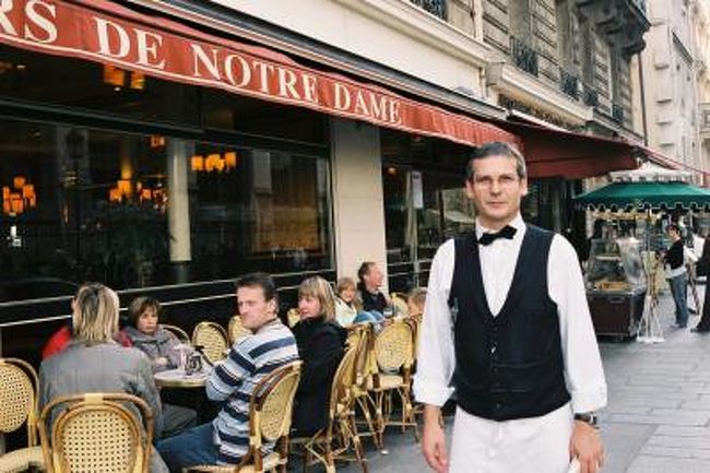 ノートルダム寺院前のカフェAUX TOURS DE NOTRE DAMEのギャルソン 2006.10.26<br /><br />はじめに<br />フランス ブルゴーニュ地方南部のボージョレ地区で造るワインの新酒、いわゆるボージョレ・ヌーボーの余韻のさめぬうちに、ぶどう畑を訪ねた旅日記を紹介します。<br />日頃のハードな業務に耐えた自分への褒美にと出かけた、写真とワイン好きを自認する私の今年の海外旅行は、いつもの9月末の夏休より1ヶ月遅れて、10月末になりました。<br />主な目的はこの時期に合わせて、ワインの名産地ブルゴーニュの、なかでも名酒 ロマネ・コンチの故郷ヴォーヌ・ロマネ村などのあるコート・ド・ニュイ地区のぶどう畑の黄葉の写真撮影でした。<br />コート・ド・ニュイ地区はブルゴーニュの中心都市ディジョンを起点に、幅約10Kmで南へ30Kmほどまでのそれほど広くない地域です。<br /><br />ブルゴーニュの前に訪れたアルザス地方の中心都市ストラスブルグは世界遺産の大聖堂、オランジュリー公園、コウノトリ、木組の家並みが美しいプティ・フランス、都市交通システム(パーク&ライド)のトラムなどが印象に残り、帰りに寄ったパリの夕焼けは、いままでこんなの見たことない、というほど感動的で、一人で見るのはもったいない風景でした。<br />(この記事は社内誌の2006年12月号に掲載したものです)<br />───────────────────────────<br />フランスの旅2006 アルザス・ブルゴーニュ・パリ<br /><br />2006年10月26日(木)晴れ パリ(最終日)<br /> 今朝はセーヌ川沿いに東へ歩きポンヌフからさらにその先のノートルダム寺院へ向かった。太陽はまだ雲間から出ず、さすがに観光客もまばらだった。寺院の裏側の庭は犬が入園禁止で入り口の扉の工夫には感心した。ヒンジの上下の軸がオフセットしていて、扉の重さでモーメントを発生させ自動的に閉まるようになっている。気にして見るとどこもそうなっていた。元祖自閉扉<br /><br /> ノートルダム寺院に入り、休憩を兼ねてオーディオガイドを聞きながら、ステンドグラスのあまりの美しさにしばし見入った。この頃には日が出て東側の祭壇のステンドグラスは直射日光を受けてまぶしいほどだった。寺院北側のカフェ・ノートルダムでコーヒーを飲みながら外を見ると、観光客がだいぶ増えてきていた。<br /><br /> セーヌの川岸伝いに歩いき、一番新しい橋ポンヌフをくぐってルーブル美術館へ。<br />昨年読んだ話題作ダヴィンチ・コードを思い出しながら、逆さのピラミッドやモナリザなどしばらくぶりに対面。ミロのビーナスもそうだ。 <br />ルーブル美術館はあまりにも大きくて覚悟して行かないと目当てのものが見られないので、今回は短時間でひき上げた。ホテルへの帰り道にセーヌ川沿いのブキニスト(屋台風の古本屋)でみやげに頼まれていたきのこの細密画を3枚20ユーロで買った。<br /><br /> 一旦ホテルへ戻り、今日も最善の方法(入浴と昼寝)で一休みしてから夕方の散策に出た。サン・ジェルマン通りを横切りモンパルナス・タワーの見える南へ行くとブランドショップがずらりと並んでいるが関心なし。<br />交差点近くのバス停のベンチに座っていると、子供連れや帰りのいろいろな人が、バスに乗り込んだり、話したりする様子が(意味はわからないが)雑踏の中のアクセントになって面白い。<br /> <br /> 旅行最後の夜は軽く日本食をと思い、ガイドブックでホテルの近くに円yenというそばやを見つけた。<br />サン・ジェルマン通りからカフェ・フロールの横を入ってすぐのところにあった。<br />前菜のマグロとアボガドのサラダは、わさび味噌との合わせ味が絶妙で白ワインもよく合う。<br />そばは鴨南蛮。やはりフランスは鴨がおいしいので。白ワインをもう一杯にデザートはアイスクリームの2色盛り。この店のオーナーは30代の日本人で、半々の従業員のサービスも行き届いていて感じよかった。<br /><br />周辺のレストランもこの時間、9時頃になると、どこも大盛況で、朝が遅い分だけ夜の始まりが遅いのかと思った。<br />一週間の旅が無事に終わり、「ブルゴーニュの葡萄畑の黄葉の撮影」という目的達成の充実感と出会った多くの人々への感謝の気持ちでいっぱいだった。<br /><br /><おわりに><br />今回の旅行の主旨は写真を撮ることでしたが、すごく運がよく、一日おきに変わる天気が私の行動にあわせて好転(好天)してくれました。<br />一日ずれたらと思うとゾッとします。<br />旅日記の内容は写真集の解説のようになってしまいましたが、今回旅ブログを作成するに当たって、このような形式の旅日記が大変役に立った。 <br /><br />撮影 CONTAX RX <br />Vario-Sonnar 3.4/35-70, Distagon 2.8/25, Planer 1.4/50<br />FUJI COLOR Pro400(ネガフィルム)<br />画像サイズ1840×1232にデジタル・データ化<br />気になる写真は元画像に拡大してご覧ください。無料です。(笑)<br /> <br />編集・公開 2010.9.23<br /><br />おことわり<br />旅行記の本文に記述の写真の一部は、編集の都合でパリその1に掲載しています。<br /><br />クチコミ「スリの被害に遭わないための予防策」2011.3.28<br />http://4travel.jp/traveler/810766/tips/<br /><br />お気に入りブログ投票(クリック)お願い<br />http://blog.with2.net/link.php?1581210<br />