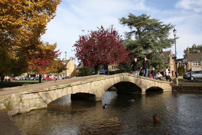 ボートン・オン・ザ・ウォーターBourton on-the-waterの石橋Stone Bridge <br /> 2010.10.30<br /><br />はじめに<br />毎年1回出かけている海外旅行は一人旅の撮影旅行が多いのですが、今年は3年ぶりに家族で行くことになりました。行き先は家族の希望にしたがって、ヴィクトリア・アルバータ博物館見学とアフタヌーン・ティーを楽しむためにロンドンと初めてのルーブル美術館見学にパリです。<br /><br />2010年10月30日コッツウォルズ日帰り観光 <br />今回の旅行はロンドンとパリが主体でしたが、イギリスの自然の風景にも触れたいと思い、最近人気のあるコッツウォルズ日帰り観光に参加しました。<br />マイバスロンドンの日本語ツアーをネットで調べ、第一希望のハイブリーが催行日でなかったのでここを選びました。<br />私自身はイギリスの自動車メーカー ローバーとの共同事業でトランスミッション部門があるバーミンガムやオクスフォードをたびたび訪れ、テスト走行でコッツウォルズを何回か走ったので懐かしい地域です。<br /><br />今回訪れたのはバーフォードBurford、ボートン・オン・ザ・ウォーターBourton on-the-water<br />、ブロードウェイBroardwayとストウ・オン・ザ・ウォルズStow on the Woldです。<br /><br />朝7:45にピカデリー・サーカスにある三越前に集合であったが、地下鉄セントラル・ラインが土曜日の工事で終日運休であるのを知らずにハイストリート・ケンジントン駅のホームで暫く待っていた。時間の余裕がなくなったのでオイスターカードの払い戻しの処理を急がせ、タクシーで向かった。出発の5分前に着き、どうにかセーフ。<br />連れのトイレ要望にはピカデリー・サーカス広場地下にある清潔なトイレを知っていて良かった。<br /><br />大型バスでガイドを含めて21名。運転手はマイバスの白髭マイクさん。5年ぶりの再会です。<br />ロンドン市内から西北西90Kmにあるオクスフォードに向かい国道40号線(A40、途中は高速M40)を走る。ゆるやかな丘陵地帯が続く。畑や牧草地帯はうっすらと緑に覆われる。<br />落葉樹は様々な色合いに黄葉・紅葉し始めているのが秋を感じさせてくれる。<br /><br />オクスフォードから更に西北西約30Km、最初に訪れたコッツウォルズの町はバーフォードBurford.<br />ford とは「川の浅瀬」とガイドの説明があった。<br />町の中心道路の両側に歴史ある様々な建物・住宅が並び、ロンドンの雰囲気とはまるで変わって心和む街並みの風景に出会うことができる。しかもそれらが歴史的展示物でなく、人々が現に暮らしている生活の場であるので息衝きが感じられる。<br />次はさらに北西12Kmにあるボートン・オン・ザ・ウォーターBourton on-the-water<br />町の中心を流れる川とそれに架かる橋を目玉にして町興し観光に成功した良い例だそうだ。近年急激に観光客が増えているようだ。<br />確かに幅20mほどの浅瀬のような川に鴨やカモメが遊び、夏なら水遊びができそうな澄んだ水が流れている。<br />周辺にレストランや各種のお店が沢山ありのんびり観光には適している。ここのレストランで昼食を摂った。<br /><br />3番目はブロードウェイBroardway<br />最初のバーフォードから北北西に21Km、コッツウォルズ地方の北の端にある。<br />たしかに比較的広い道路broardwayが通っている。<br />ガイドの説明では有機野菜を売る八百屋・果物屋が人気とか。店には「オーガニック・アイスクリーム」の看板があった。植物性油脂から作るのだろうか?試してはみなかった。<br />通りの裏側を歩いてみると、コッツウォルズ地方特有の黄色い石造りの真新しい家に出会った。また石造りで藁葺き屋根の家があり、その前にホンダのjazz(フィット)が停まっていた。<br /><br />最後はコッツウォルズ地方の中心地ストウ・オン・ザ・ウォルドStow-on-the-Wold<br />バーフォードの北西10Kmにあり、8つの道路が交差する交通の要所。<br />コッツウォルズ丘陵で標高が240mで最も高い。<br />ここはティータイム休憩で、石畳の広場マーケット・スクェアに面したOLD STOCKS HOTELの日当たりの良いティールームでクリーム・ティーを味わった。イメージしたのは「ミルクの代りにクリームを入れた紅茶」であったが、出されたのはスコーンとそれにつけるジャムとクロテッド・クリーム、および紅茶でミルクはたっぷり用意されていた。アフタヌーン・ティーのスコーン・セットと同じです。<br />お茶する前に同行の有志とホテルの前で記念写真を撮った。<br /><br />帰りは今朝と同じルートをオクスフォードから国道40号A40(高速M40)を通ってロンドンへ戻った。<br />夕日を浴びたあたりの風景も黄葉が美しく、秋の気配を楽しんだ一日だった。<br /><br />撮影 CANON EOS40D EF-S 17/85 IS USM, Nikon Coolpix 5400<br />yamasda423<br /><br />写真と説明文をひと通り読んだ後、元の画像に拡大してご覧いただくと,<br />文字や人の表情などがかなり詳細に見られますのでお勧めします。<br />次の写真へ>> でページが送れます。<br /><br />現地の日の出・日没時刻はこのクチコミ記事で簡単に調べられます。<br />http://4travel.jp/overseas/area/europe/france/mont_st_michael/tips/10177076/<br /><br />参考:個人で旅行される方に。2018年6月追記<br />Cotswolds Discoverer<br />コッツウォルズ地方のバスおよび鉄道の1日パス 10ポンド<br />路線図も付いています。<br />http://ukbytrain.com/cotswolds-discoverer/<br /><br /><br /><br />