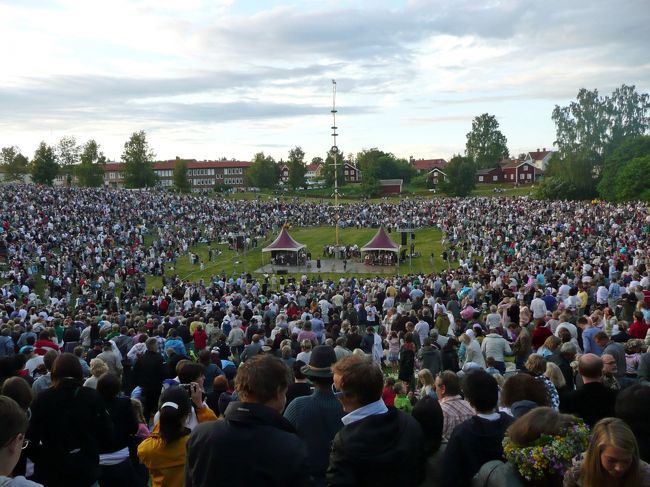 【6日目】6/25（金）<br />スウェーデンでは，夏至に最も近い土曜日（Midsommardagen）とその前日（Midsommarafton）が祝日となり，その前後に各地で夏至祭（Midsommarfirande）が行われます．夏至祭の中でも一番のイベントは夏至柱（メイポールとも言う，Midsommarstång）と呼ばれる花飾りを立てることです．大きなものでは20 mを超える大木に白樺やモミの葉や花々を美しく飾り付け，その周りで民族衣装を着た人が音楽や歌を奏で，それに合わせてみんなでフォークダンスを楽しむのが一般的な光景です．<br />　昨日もメイポールを立てるイベントがありましたが，一番盛り上がるのが今日，夏至前夜祭です．町外れにある大広場で18時頃から式典が始まり，来賓挨拶や演奏，メイポール搬入の儀式などが行われている間に，徐々に人が集まり始まり広場を埋め尽くしていきました．<br />メイポールが垂直に立った後，内側半分くらいの人々がメイポールの周りを回りながらフォークダンスを踊りました．教会でのコンサートを聴くために途中で広場を離れましたが，おそらく22時頃までイベントは続いていたと思います．もちろん太陽はまだ沈んでいません．ふだんは静かな町ですが，この日は夜中を過ぎても若者の盛り上がる声が聞こえました．