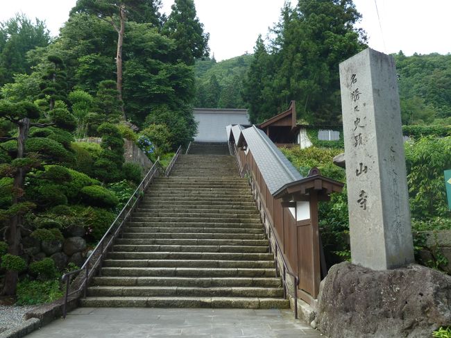 芭蕉達は、尾花沢滞在中のぐずついた天候がやっと晴れたのを見計らって、当初の予定には無かったが、尾花沢の人々の勧めに従って、やや後戻りする道順になるのも構わず立石寺を目指す。<br /><br />清風が用意してくれた馬で楯岡まで行き、その後は徒歩で南東に向けて進み、天童に入り、一日町の辺りから山寺街道を西に辿り、山寺の宿坊に辿り着く。<br /><br />芭蕉達は宿坊に荷物を預け、その日の内に山寺の巡礼を終える。<br /><br />尾花沢を発った我々のバスが立石寺山門に到着したときはまだ小雨が降っていた。<br /><br />しかし、1015段あるという長い石段を登っているうちに、いつに間にか雨は上がり、奥の院から開山堂を望む頃には夏の太陽が顔を出した。<br /><br />お陰で写真の数も多くなり、<br />　①参道から見上げた宝珠山と点在する堂宇と山門まで、<br />　②山門を潜り、800段の石段を登って奥の院下まで、<br />　③奥の院前から開山堂、五大堂のパノラマ<br />の3回に整理して投稿します。<br /><br />正式名称は宝珠山立石寺だが、立石寺という堂宇があるのではなく、山寺同様全山の総称。<br /><br />我々が目にする土産物屋や飲食店並ぶ門前の通りは、江戸時代宿坊が立ち並んでいたらしい。<br /><br />山を降りてから気がついたのだが、その門前通りから見上げると、宝珠山が聳え、良く見ると山を覆う樹木の間に荒々し岩肌がが覗き、その上に五大堂や開山堂の姿がくっきりと見える。<br /><br />更に目を凝らすと赤い納経堂までも望める。<br /><br />その門前の通りに沿って石段の片方だけが築地塀を持つ、端正な山寺の入り口はある。<br /><br />石段を登ると正面に、宝珠山立石寺の開祖、慈覚大師を祀った、入母屋造りの根本中堂が現れる。<br /><br />根本中堂の左奥に鳥居が立ち、その先に、珍しく芭蕉と曾良が離れて置かれた銅像がある。<br /><br />念仏堂と鐘楼が並んで立つ一角を過ぎると「開北霊窟」と書かれた扁額を掲げた山門前に出る。<br /><br />根本中堂とこの山門だけが、山寺の幾多の火災を免れ、創建当初の姿を残しているとのこと。<br /><br />山門前をそのまま真っ直ぐに進むと、2009年ここを訪ねた帰りに潜った「抜苦門」があり、立石寺本堂の屋根も見える。<br /><br />此処に入場券の発売所がある。<br />