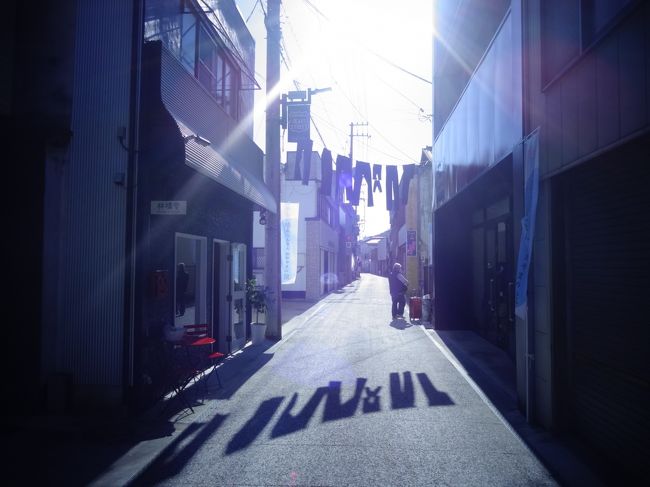 <br />海に近い一山越えた山間の街の隠れ家のような鮨屋でたらふく美味い鮨を食って、帰りは児島駅から一駅の上の町駅であるが、そこは普通電車しかなく、しかも特急との接続が驚くばかり悪いのでタクシーで児島駅まで行くとことにする。<br />勘定を済ませ大将に再訪を告げて店を出ると、鮨屋の女将さんはタクシーに乗り込んだ私に向かって深々とお辞儀をしていた。<br />手を振り別れ、ちょっと昼からしこたまお神酒が入っていたのでいい気分になっておりタクシーのなかで妙案が浮かぶ。<br />せっかくめったに来れそうもない場所へ来たのだ。<br />児島ジーンズストリートや旧野崎家も観光してみるべきではないか。<br />「旧野崎家って見所あります?」<br />そういえば、鮨屋の店主には苦笑いで何も返事もらえなかった気がするが。<br />丁寧な初老の運転手さんに行先を旧野崎家への変更を告げる。<br />先週児島駅構内で見かけた観光案内看板では、児島駅と上の町駅とのちょうど中間にある。<br />ついでにジーンズの聖地、と謳われるジーンズストリートも歩いてみようではないか。<br />そこから駅まで歩けばいいや。<br />旧野崎家の正門前で降ろしてもらう。<br />「ここからジーンズストリートは遠いんですか?」<br />「いえ、この通りからもうそうですよ。ほら、その建物が桃太郎ジーンズの本店ですよ」<br />なんだ、ますますここに来てよかった(笑)。<br />野崎家巡りもジーンズストリートも秋晴れのすばらしい陽だまりのなか楽しめた。<br />通りに吊るしてあるジーンズに一番陽が高い時間帯で真下に影が映る。<br />福山の「思い出横丁」のような、しかしこちらは本物の生きた昭和の匂いがした。<br />地元の通りすがりのおばあさんが「撮ってばかりじゃなくて写らなくていいの」とカメラマンを申し出てくれ、ますます昭和の味がする。<br />ここ、児島も今では行政区は倉敷市。<br />ジーンズの影がますます倉敷のことが好きなった自分の影法師にも見えた。<br />好きだな〜倉敷。<br />高知の長女といい、倉敷の二女といい、親思いの娘さんたちだ(笑)。<br />ジーンズストリートという名称のほうがすっかり定着した野味商店街を過ぎると、倉敷児島交流センターがあり、そこの広場がなかなか風情があってよろしい。<br />あとで知った、この野味公園は通称橋公園、瀬戸大橋開通を記念して整備された公園らしい。バブル前夜の歴史を感じる。<br />全国の有名な11の橋を模した回遊公園らしい。その昔はすべて水辺だったがいまでは砂利が敷かれていて時代の侘しさを感じさせられる。<br />そこでしばらく遊んだあと(本当にアスレチックコースみたいなものもあって)海側へ歩を進めていくうちに近くからスティームドラムのような音とスピーカーから流れるリズム感ある男性の声が響いてきた。中近東や北アフリカで何度も何度も遭遇したお祝いごとのあのリズムだ。<br />ピラミッドのすぐ袂にある迷路のようなナズラット・サンマーン村に悪徳ラクダ使いに誘われたと結婚式のときの光景が浮かぶ。<br />あのときも一目散に角を曲がった。<br />ヴェネツィアのサンマルコ広場でも海上タクシーを降りた途端、ハドラマウト砂漠を越えたイエメンのサユーンの旧市街の広場でも、マラケシュのジャマエルフナ広場でも、カメルーンの首都ドゥアラの空港ビルをでたときにも、いつもわくわくするような音があった。<br />ワクワクする音に誘われ一目散に走っていた。<br />素敵な出会いが待ち受けている予感がする音楽、ドラムの音。<br />これはすぐにサンバだとわかった。児島の鷲羽山ハイランドという遊園地はサンバの国としても売りである。その影響だろうか、街中でもサンバ?<br />小走りで音の方に進むと、そこは駅前の商店街でふたりのブラジル人女性が例の寒々しい衣装でドラムのリズムに合わせて踊っていた。そして周りを取り囲むのはこの季節になるとテレビで最近よく見かけるような思い思いの衣装に身を包んだ市民たち。<br />こじんまりした野外ハローウィンパーティーをしている様相。<br />秋晴の空の下、ドラムの音につられいつまでもここにいたい心地がしたが、当初の予定より一列車遅らせていることに気を取り直して離れることに。<br />陽は随分西日となっていた。<br />ジーンズストリートでみた影とは明らかに違っていた。<br />児島駅のホームからでもドラムの音は風にとどき、列車に乗り込んでもいつまでも耳に残った。<br />瀬戸大橋を渡った途端、その音色がかき消された気がした。<br />影法師と戯れた一日だった――――。<br /><br /><br /><br /><br /><br />★「ジーンズの聖地 児島ジーンズストリート」<br />児島地区は、明治時代から「繊維の町」として知られていますが、ジーンズに関しては1960年代に国内で最初にジーンズ生産を手掛けたことでも有名です。<br />この「児島ジーンズストリート」は、地元メーカーや児島商工会議所等による協議会が味野商店街の空き店舗への誘致活動として行っております。<br />「児島ジーンズストリート」と名付けられたその通りは、かつて児島で最も栄えた味野商店街の中にあり、旧野?・家住宅前から味野第2公園までの400メートル程のストリートとなっています。<br /><br />★児島ハロウィン・ホワイト<br />このイベントは児島駅前商店街連盟とのコラボ企画でありハロウィンとKOJIMA WHITE(イルミネーション)の点灯式を協働して行うイベントです。<br />ハロウィンでは商店街へお菓子をもらいながらパレードしたり<br />豪華景品のあたる抽選会、ランタン作り、仮想コンテストなど楽しい事が盛りだくさんありますので、家族、友人を誘ってご参加ください。<br />13:00〜受付開始 (ランタン作り 先着50名) <br />14:00〜パレード開始ですので、お早目にお越しください。<br />また、陽が落ちるとKOJIMA WHITEの点灯や2000個のキャンドルが点火し、会場が幻想的な灯りに包まれます。詳しくはFacebookでもUPしておりますのでご覧ください。<br />この週末は仮装して、ぜひ児島へ!<br /><br />★旧野崎家住宅<br />野崎家は、製塩業と新田開発で財をなした野崎武左衛門がその気宇に合わせて天保から嘉永年間に次々と築いていった民家である。<br />敷地面積は約3000坪・建物延床面積は、1000坪近くある。<br /> 武左衛門は、文政10年(1827年)から文久3年(1863年)にかけて、野崎浜(現在の児島駅附近)、日比亀浜、東野崎浜、久々井浜<br />など終生161ヘクタールの入浜式塩田を開発した。<br /> その間、岡山藩の命によって、福田新田652ヘクタールの大干拓事業も完成させた。その謹功により、嘉永6年(1853年)に大庄屋に取り立てられた。<br /> 武左衛門の全盛期に次々と建てられたこの建物は、創建当時の建築技術の粋をこらしたもので、独特な平面の主屋、意匠に優れた表書院など<br />庭園と共にそのまま今日に伝えられており、雄大な規模を有する当時の大事業家の居宅として貴重である。(財団法人 竜王会館)<br /><br /><br />