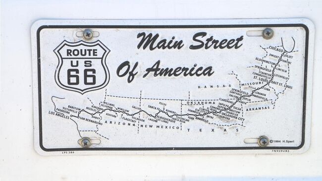 Route 66 は、1926年に造られたイリノイ州シカゴと、カリフォルニア州サンタモニカを結ぶ全長3,755km（2,347マイル）の、アメリカ合衆国をほぼ横断する国道。スタインベックの「怒りの葡萄」では The Mother Road 、国道66号線協会による「公式」な別名としては The Will Rogers Highway 、プロモーションでは The Main Street of America と呼ばれた重要な国道だった。<br /><br />しかし、WW2後、敗戦国ドイツでアウトバーンを目の当たりにしたアイゼンハワー大統領が国策で整備した インター・ステート（州間高速道路）の完成によりその役目を終え、1985年に廃線となった。その後、1990年にアリゾナ州とミズーリ州で存続運動が起こり、それぞれルート66協会が発足、同年、ミズーリ州政府は州内の国道66号線を州の歴史的街道に指定すると宣言した。<br /><br />今回のツアーで立ち寄ったセリグマンという町もR66の存続運動では重要な役目を果たした町。セリグマンで床屋を営んでいたエンジェル・デルガディーロと友人達が「Histric Route 66 Association Arizona」を立ち上げ、Arizona の議会に、歴史的なルート66の保存を認めるよう働きかけた。やがて運動が実り、歴史的な道・ルート66は、再び地図上に名前が刻まれるようになった。現在I・Sに組み込まれてしまった部分を除き、旧R66の80%ほどが実際に走行できるそうだが、人間が関係する古い物が少ないアメリカでは貴重な歴史的遺産となっている。<br /><br />写真は今回乗車したバスのナンバープレートの隣に取り付けてあった The Main Street of America のプレート。まあ、日本の「東海道六十三次」みたいなものだ。なお、この66という数字は、覚えやすく、言いやすく、聞きやすいという理由でゾロ目の番号66があてられたとのことだ。<br /><br />2024/03/06　一部修正