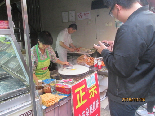 中国人は朝食、昼食、夕食の三食を外食してます。古い住宅には台所がありません。子供のお弁当も作りません、お店で買って持って行きます。サラリーマンも朝食を買って会社に持って行きます。朝ご飯を食べ終わらないと仕事をしないとか言われています。定番の朝食は肉まん、野菜まん、ロボス（切干大根入り）、お粥、油条（揚げパン）豆乳などが代表的で種類も沢山あります。以前は電車やバスの中で食べていましたが現在は禁止となっています。朝は６時頃から営業しています。今でも移動車、屋台が来て販売していますが、屋台は取締りの対称に成っています。店舗も営業許可を持って居ない店、保険所の許可の無い店はビシビシ取り締まりを行っています。マクドやＫＦＣで朝食セットでお粥を販売しています。ここ広西北路に7軒集まっています。毎朝同じ物を食べずに済みます。店に寄って味も違うでしょう。選ぶ楽しさが必要でしょう。大勢の人が集まる上海らしい景色です。