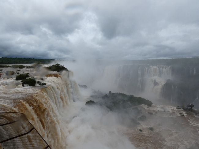 12時15分過ぎ、イグアスの滝ブラジル側の散策を開始。地理的には滝のうちブラジル領となるのは、悪魔の喉笛(Garganta do Diabo)で有名なウニオン滝(Salto Union)のブラジル側手前(右岸)寄りのフロリアーノ滝(Salto Floriano)など、全体の20%程度なのだが、左岸に広がる残り80%の部分も含めて全体が把握できる点ではアルゼンチン側より優れている。確かに最初に川が見えた時に見た流れ落ちる滝群には言葉が出なかった。いや、なんちゅう数になんちゅう水量。この時にはまだ把握できてなかったが、最初に見えたこの部分は翌日訪ねたアルゼンチン側のアッパートレイル(Circito Superior)やロウワートレイル(Circito Inferior)の終点辺りで、川がUターンしているので川幅が広がっているが、その回り込んでいる一番外側の部分で、その左手にあるのは滝に削られずに残っているサンマルティン島(Isla San Martin)だった。凄いとしか云いようがない・・・<br /><br />しばらく滝に感激した後、川の右岸に続く遊歩道を上流に向かう。ここから1.2㎞ほど遊歩道が続く。10分も行かないうちにサンマルティン島の上流側に抜けるが、ここは滝が2段になっており、奥に続く水のベールと、すぐ向かいの2段目の滝群を見通せる展望台がある。私も並んで写真を撮ってもらう。すぐ向かいにある2段目の滝は三銃士の滝(Salto Tres Mosqueteiros)と云い、上の段のリバダビア滝(Salto Rivadavia)から続いている。雨は上がったのだが、水しぶきがすごい。ここから上流の悪魔の喉笛方向が見えるのだが、先は水しぶきで見通せない。三銃士の滝を過ぎたところにも展望台がありそこでも記念撮影(下の写真1)。<br /><br />この遊歩道、たくさんハナグマ(Coati)がいることいること。正確にはアカハナグマ(South American Coati)で、南米の中央部の森林、低地の草原地帯に棲息しているアライグマ科の動物。アライグマ(Raccoon)同様、見た目は可愛いが結構狂暴と云うことで手を伸ばすようなことはしなかったが、確かに可愛い。以後、イグアスの滝ではアルゼンチン側も含めて山ほど見掛けた。あと、オオトカゲ(Monitor lizard)もたくさんいた。<br /><br />ゆっくり写真を撮りながら歩いたので30分ほど掛かって、一番奥の滝に到着。これがフロリアーノ滝(下の写真2)。目の前を泥水が大量に流れ落ちる。声が出ない。このフロリアーノ滝も2段になっており、2段目のサンタマリア滝(Salto Santa Maria)の上を悪魔の喉笛の手前まで遊歩道が繋がっている。しっかり防水していよいよ先端に向かう。足元を通った水がサンタマリア滝を流れ落ちるのも凄い。で、一番先端、悪魔の喉笛のウニオン滝の滝壺が見えるところまで進んだが、凄い水しぶきで正直あまり何も見えない(下の写真3)。ウニオン滝は約50mの落差の1段滝なので、一気に流れ落ちてる。<br /><br />フロリアーノ滝の袂まで戻り、エレベータで上階に上がると、そこからの景色もまた少し違って見えてなかなか面白い。2時前になったので、滝の上部部分にある休憩所ポルトカノアス(Porto Canoas)のファーストフードでハンバーガーのお昼とする(下の写真4)。29.99R$だったので1100円くらい。この休憩所からは滝の手前の部分が見える。アルゼンチン側の悪魔の咽笛手前の遊歩道の先端に人が溢れているのもよく見える(下の写真5)。<br />https://www.facebook.com/chifuyu.kuribayashi/media_set?set=a.1874176532652362&amp;type=1&amp;l=8a89379cb0<br />https://www.facebook.com/chifuyu.kuribayashi/videos/vb.100001801017376/1874305405972808/?type=1&amp;l=8a89379cb0<br /><br />その後再度遊歩道を歩いたりして3時半頃にポルトカノアスからマクコサファリ(Macuco Safari)へバスで移動し、ボートツアーでイグアス川(Rio Iguacu)を上る。ツアーは215.4R$(約7500円)とちょっと高いけど、しゃあない。チケットを買ってまずはグループになって電気自動車で移動、その後ボート乗り場まで10分ほどジャングルを散策。濡れてもいいように水着に着替えてボートに乗船。ここからまずは今日最初に滝が見えたサンマルティン島のところまで川を遡る。これが10分ほど掛かる。ちょうどここら辺りで晴れてくる。ここでタイミング待ちを5分くらいして、ボートはいよいよ滝に突入。この時には分かってなかったが、ブラジル側のボートツアーで突入するのは遊歩道から見てた三銃士の滝。水の下に何回も突っ込んでくれる。まあ、なかなか面白い体験だった。ちなみにアルゼンチン側のボートはタイミング待をしてた辺り、ローワートレイルのサンマルティン島の向かいからボートに乗り込み、奥のサンマルティン滝(Salto San Martin)に突っ込むようだ。<br />https://www.facebook.com/chifuyu.kuribayashi/media_set?set=a.1874194342650581&amp;type=1&amp;l=8a89379cb0<br /><br />5時15分頃ボート乗り場に戻り、本日の観光は終了。再度着替えて電気自動車でバス道に戻り、バスでゲートに戻り、路線バスで町に戻る。ホテルには7時頃戻る。翌日の朝、アルゼンチンの移動、ここに来るまではバスで国境へ移動し、いったんバスを降りて出国し、次のバスでアルゼンチン側に橋を渡り、またバスを降りて入国審査して、三度バスを待ってアルゼンチン側の町に移動しようと思っていたのだが、大きなスーツケースを持ってのこの移動は無理と判断。アルゼンチン側のホテルまでへのタクシーを頼むことにする。ところが、朝には英語しゃべれる兄ちゃんがいたのだが、ホテルのフロント、誰も英語が喋れない。結局、その兄ちゃんに電話してくれて、ようやくOK。ああ、しんど。まあ、でもこういうシチュエーションは40年前に初めてヨーロッパに旅行した時から何度も経験しており、なんとかなるもんだ。<br /><br />少しンホテルで休んだのち、8時頃から夕食。ブラジル最後の夜と云うことで、再びシュラスコ(Churrasco)を食べに、10分ほど歩いてシュハスカリアトロピカーナ(Churrascaria e Pizzaria Tropicana)へ。たっぷり食べたわ。ビール飲んで、ワイン飲んで、90R$(3000円足らず)だったので、満足! 帰り道で道端で売ってたTシャツを購入して9時過ぎにホテルに戻った。この日は2万1千歩越えで、ブラジルではサンパウロ、リオと併せて毎日よく歩いた。4日間で10万歩以上。<br />https://www.facebook.com/chifuyu.kuribayashi/media_set?set=a.1874203539316328&amp;type=1&amp;l=8a89379cb0<br /><br /><br />イグアスの滝、アルゼンチン編へ続く。