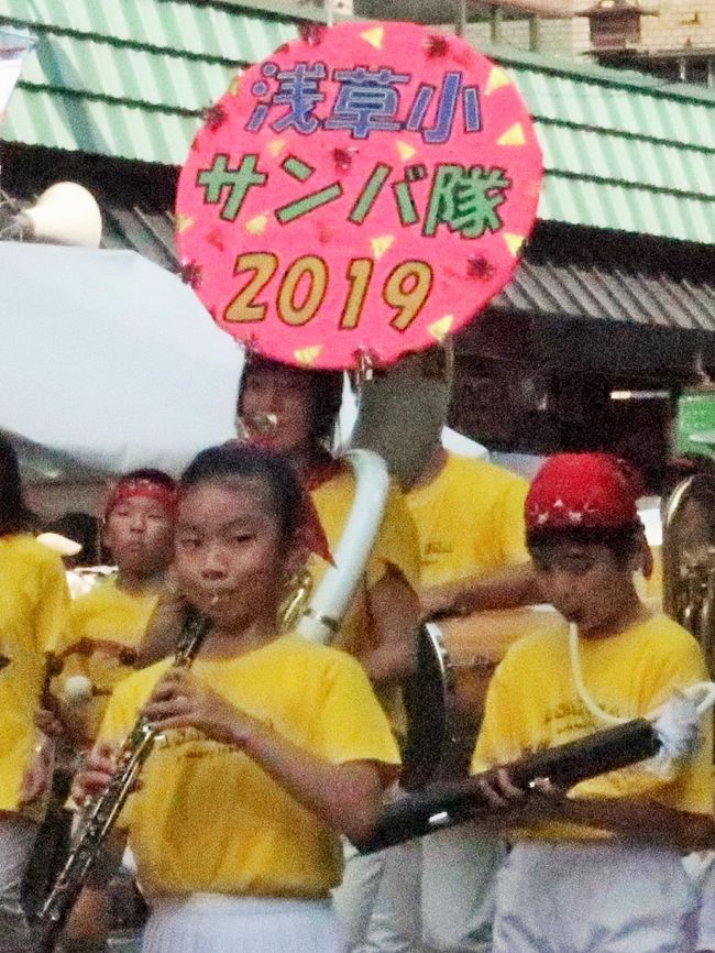 先陣はつらつ浅草っ子行進　浅草サンバカーニバル<br />　今年もカーニバルの先陣は「浅草っ子」たちだった。コンテストの前のコミュニケーションリーグでは、区立田原小（雷門一）の田原バンド・フラッグ隊、区立浅草小（花川戸一）のサンバチームが練り歩いた。<br />　桃色のＴシャツの田原小は4～6年総勢130人ほど。ブラスバンドの演奏に合わせ、青と黄色の間にピンクで「ＴＡＷＡＲＡ」と記された旗を華麗に回し、沿道の観客を沸かせた。<br />　背中に「令和」の文字と干支（えと）のイノシシをあしらった黄色のＴシャツの浅草小は、児童に保護者らも加わり150人ほど。ブラスバンドの「ダイヤモンド」などの曲に合わせて、「イェーイ」とカラフルなボンボンを振り、元気いっぱいに行進した。(東京新聞より引用)<br />