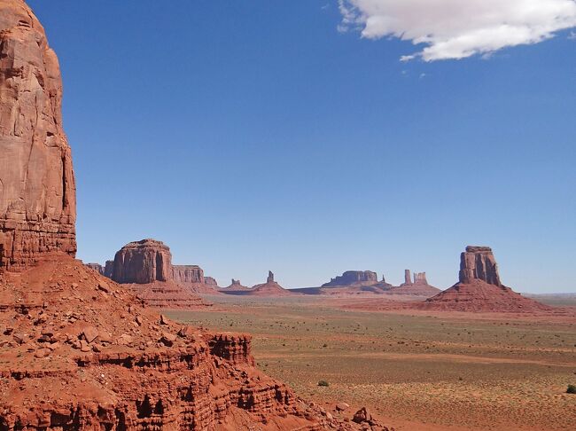 風化・浸食によって形成された最高300 mにもおよぶモニュメント状の岩が、広大な赤茶けた平地に点在するモニュメントバレー（Monument Valley）を見学して来ました。本旅行記は「アメリカ　グランドサークル　メサ・ヴェルデ国立公園　フォー・コーナーズ」（https://4travel.jp/travelogue/11696738）の続きです。このバレーはナバホ・ネイション（Navajo Nation、ナバホ族の準自治領（インディアン保留地））内にあって、フォー・コーナーズの西約80 kmのユタ州とコロラド州にまたがった位置にあります。モニュメントバレーは公式には面積約370 km^2（東京23区の1/2強）のモニュメントバレーナバホ部族公園（Monument Valley Navajo Tribal Park）を含む広大な領域を指しますが、一般にモニュメントバレーというと、後者を意味するようです。 　<br />モニュメント状の岩は一般に頂上が水平で側面が鉛直状になっていますが、地質学者は経験的に頂上の幅が高さより広い場合をメサ（mesa、西語で「テーブル」の意）、狭い場合をビュート（butte、仏語で「小さい丘」の意）と呼んでいます。ビュートやメサには名前がありますが、これはナバホ族によるものか最初の開拓民がその形から連想されるものの名前を付けたものです。　<br />モニュメントバレーではジョン・フォード（John Ford）監督の西部劇を含めたくさんの映画が作成されており、私たちが宿泊したホテル横にはこれら映画関係の資料等を展示したミュージアムがあります。本旅行記では前半でこのミュージアム、後半でモニュメントバレー内のバスツアーについてご報告します。<br /><br />付録に、モニュメントバレーの１．地理、２．地質、ここで撮影された、３．映画のあらすじについて記載しましたので、お時間とご興味のある方はご覧ください。<br />
