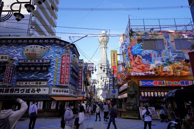 大阪市浪速区恵美須東にある新世界は、ビリケンさんで有名な通天閣をはじめ、色とりどりの看板があふれる商店街など、コテコテの大阪が満喫できる超人気の観光スポットです。<br /><br />明治３６年（１９０３年）に開催された第５回内国勧業博覧会の会場跡地の西側に、パリのエッフェル塔をモデルにした初代通天閣が明治４５年（１９１２年）に建てられたことを契機に、芝居小屋や映画館などが集まる、賑わいのある新世界がかたちづくられていきました。<br />（ちなみに東側には天王寺公園が整備されました。）<br /><br />その後、火災に見舞われた初代通天閣は解体されてしまいましたが、『なにわのシンボルの復活を』との市民の熱い要請を受けて、昭和３１年（１９５６年）に２代目の通天閣が再建され、大阪を一望できる展望台や、足の裏をなでると幸運が訪れるというビリケン像などが人気を呼び、年間１００万人以上の観光客が訪れています。<br /><br />なお、新型コロナウイルスの感染状況を府民に周知するため、「大阪モデルの指標」となる赤・黄・緑の信号を通天閣に点灯しているのはご存じの通りです。<br /><br />このコロナ禍にもかかわらず、芝居小屋や映画館をはじめとする娯楽施設、たこ焼きや串カツなどの安くて美味い大阪グルメなど、「これぞ大阪！」という雰囲気を求めて、今日も人々が集まっています。