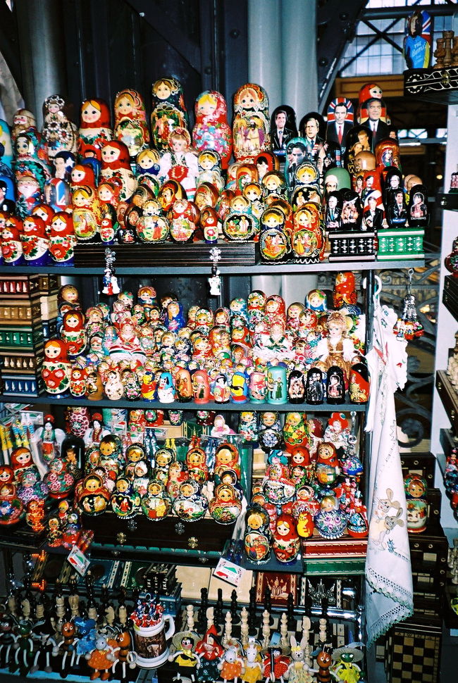 2階のみやげ屋を見て回りました。カロチャ刺繍と、それからマトリョーシュカが特に目を引きました。マトリョーシュカは、写真のような展示の仕方が多く、中から小さい人形を出すディスプレイをしている店は、ブダペストのほかの土産屋でも（それからこのあとに回ったチェコでも）、私が見かけた限りでは、ほとんどみかけませんでした。これに対してロシアのみやげ屋では、たいてい、中の小さい人形を出して、大きさ順にずらっと並べていて、見事でした。しかし、この写真のようなディスプレスも、こけしのような優しいフォルムでカラフルで可愛い人形がぎっしり並んでいて、それだけでなかなか壮観です。よく見ると、ここでも政治家の顔をした風刺のマトリョーシュカなどがあったりします。<br /><br />カロチャ刺繍は、ここのが特に割安というわけではありませんが、それでも繁華街ヴァーツィ通りで買うよりは、多少、安いらしいです。せっかくだから買うことにしました。でも、どれもステキで、目移りして仕方ありません！　そして同じような図柄でありながら、よく見るとみんな少しずつ違うのです。なので、パプリカの図柄があるものにこだわってみました。<br /><br />それでも迷う私に、お店の人は、黒い布に刺繍が施された新しいタイプのものはどうか、と薦めてくれました。けれど、ハンガリー初めて、カロチャ初めての私の目は、白地に赤い花がメインのオーソドックスな図柄に引き寄せられます。黒地のものも、とってもシックで上品でステキなのですが、なんだか上級コースってかんじがしたのです。手を出すのは、もうちょっとカロチャを極めてから―――というか、何枚か持ったあとでいいと思いました。それにオーソドックスな図柄でないと、いかにもハンガリーみやげって主張しづらそうです。伝統的な刺繍というのは、それこそ世界全土にありますから。そんな私に、店の人たちは「日本人は、みんなこの図柄が好きね」と口々に言いました。たぶん、たいていの人は私と同じようなことを考えたからではないでしょうか。<br /><br />食料品の屋台が並ぶ1階も回ろうと思って階段を下りかけたら―――なるほど、このアングルなら、いいです。市場全体を見下ろせるし、プレハブ屋根ばかり目立たなくて、なかなかよいではないですか！　写真を撮りました。（写真は、「2004年夏のブダペスト・ウィーン・チェコ旅行20日間　旅程一覧＆ハイライト写真(1)ハンガリー編」をご参照ください。）<br /><br />1階の店は、店先に干しパプリカや玉ねぎが千羽鶴のように吊り下げられているのがなかなか面白かったです。香辛料のお店はトルコやチュニジアの市場を思い出します。でもこの市場では、量り売りよりも、きれいに袋づめされているのが多いようでした。野菜売り場も面白いです。パプリカといえば、赤いとうがらしのような形のものかと思っていましたが、黄色っぽくて、特大サイズのピーマンのような形のパプリカもたくさん見かけました。2階の店もそうでしたが、1階の店も、どれも写真に撮りたくなって、きりがありませんでした。<br /><br />最後に地下も覗いてみました。日用雑貨の店やちょっとした代理店が並んでいるようでした。地上階がとても面白かったので、あんまりパッとせず、とりたてて興味を引きませんでした。客も少なかったです。ただし、おかげでベンチは空いていて、一休みするにはちょうどよかったです。