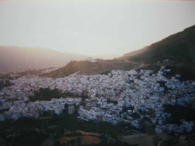 Sunset in Chefchaouen<br /><br />スペインからのフェリーが着いた港町はタンジェ(Tangier)<br />バスでシャウエン(Chefchaouen)へ。<br /><br />ヨーロッパから渡ってくる人々が最初に来る町、タンジェ。<br /><br />フェリーを降りるとすぐに<br />「政府のオフィシャルガイドだ」<br />「私たちは外国人観光客を守るためにいる」<br />ギラギラした目の男たちがわんさか寄って来る。<br /><br />駅に行くんだ、と告げると<br />「電車は遅れている。後で連れてってやるから」<br />と、別のどこかへ連れて行こうとする。<br /><br />ヨーロッパしか旅をしたことのないあたしにとって<br />モロッコ人の強引さは衝撃的だった。<br />親切なのか　それともビジネスなのか<br />真実なのか　それともデタラメなのか<br />さっぱり分からない。<br />怪しい感じはするけれど、何を疑えばいいのかも分からない。<br />相手はプロ、こちらは素人。<br />初試合に負けても仕方ない。それは勉強。<br /><br />バスの乗り場でおとなしくバスを待つ。<br />人々の好奇の目。<br />暑い。<br /><br />やっと来たバスはオンボロ。<br /><br /><br />シャウエンはタンジェとは違い、静かで小さな町。<br />バスを降りて町の空気を吸う。ホッとした。<br /><br />ホテルに荷物を置いてバザールを歩く。<br />知り合ったのはモロッコ人Mobarek。観光客用の店を出している。<br />彼の店に座り、お茶を飲みながら話をする。<br />彼は上手に英語を話す。フランス語はもっと上手い。<br />でもあたしは彼のことを知らないし、彼はあたしのことを知らない。<br /><br />あたしの警戒心を察した彼はどこかに電話をかけだした。<br /><br />彼が生まれ育ったのはシャウエンからずっと南の砂漠の町。<br />その彼の実家にしばらく滞在していた日本人女性がいるという。<br />現在はどこか違う町に住んでいる彼女。今でも家族の一員のようにつき合っているらしい。<br /><br />Mobarekから電話を渡され、彼女と少し会話をした。<br />お互いに何を話していいのか分からない。<br />でも彼女の日本語を聞いて、なんとなく安心した。<br /><br />彼には店を共同で経営している相棒がいる。<br />その相棒は「関根勤」にそっくりで、日本人の奥さんと小さな子供が居ると言う。<br />写真を見て笑った。ぽっちゃりした関根勤。<br /><br /><br />Mobarekと友達になってシャウエンの町で過ごした時間は<br />「旅行で訪れている」というより<br />「暮らしている」に近い感覚だった<br /><br />朝の強い日差しがシャウエンのまっ白い壁に反射すると<br />目も開けられないほど眩しい。<br /><br />牛や羊のフンがそこらじゅうに転がる狭い路地。<br />すれ違う人と笑顔で挨拶する。<br />丘の上から見る夕日。<br />シャウエンの町に夜が訪れるしるし。<br /><br />居心地の良い町だった。