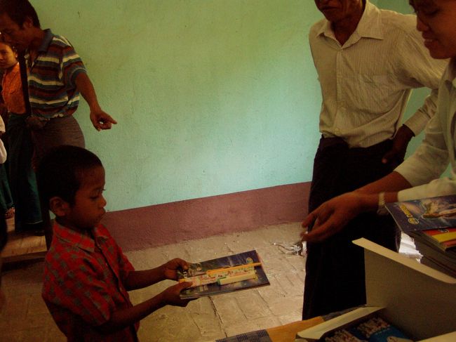 　僕と女の子が文房具を揃えてウィンウィンに渡し、彼女が生徒たちに渡すということになった。鉛筆を箱から取り出し、５本数え、女の子が持つノートの上に置き．．．と繰り返していると、数分で汗だくになった。生徒たちは一人一人、恭しく受け取っていった。時折数人の生徒がなだれ込んで来たりしたが、そのたびに先生が追い出していた。<br />　配布はスムーズに終わり、子供たちはそのまま帰っていった。何人かは、僕のことが珍しいのか、じっと僕の一挙手一投足に注目している。なんか居心地が悪い。<br />　配布が終わったあと、先生に教室の中を見せてもらった。質素な長いすと机が並んでいた。先生になにか必要なものはあるかと尋ねると、やはり椅子と机が足りないと答えた。全部揃えるのに必要な費用を尋ねると、100ドルにも満たない。今度は、机と椅子を援助してやろうか。<br />　ウィンウィンが独り言のように「前来た時は、校舎も椰子の木で作っていたけど、随分立派な校舎になってる」と言った。確かに、田舎の小学校の割にはモルタル造の立派な校舎だ。「でも、椰子の木の校舎の方が涼しいし、いいんじゃない？」「３年に一度は建て替えなきゃならないから大変よ」「でも、よくこんなお金が出たね」。ウィンウィンを通じて、先生にこの校舎はどこかの援助なのか聞いてもらった。「日本人旅行者だって」。まさか旅行者が．．．。おそらく、民間の援助団体あたりだろう。<br />　ミャンマーに興味を持つようになって数年になるが、このように学校や子供たちに援助の手を差し伸べる日本人がけっこう多いのには驚かされる。カメラマン氏もそうだし、僕がよく出入りしているミャンマー関連のサイトのオフ会に出たときも、シャン州で校舎を建てたという年配の方と会ったことがある。そして、こうした援助をしている年配の方々に共通しているのは、親、兄弟、あるいは親戚の誰かがこの地で戦死しているということだった。せめてもの罪滅ぼしと考えているのか、あるいは何らかの縁を感じているのかわからないが、彼らにとってこの地に生きる人々は、特別な存在なのだろう。<br />　校長先生に挨拶をし、車に戻った。ドライバーは「アツイ？」と日本語で聞いてきた。彼にはあちこちで随分待たせて悪いなという気持ちが強くなってきた。<br />　帰りは、配布を手伝ってくれた女の子ともう一人の幼い女の子が乗り込んできて、一緒にニャウンウーに戻ることになった。この村からはニャウンウーへの車はあまりないらしいので、これ幸いにと久しぶりに町へ行くのを楽しんでいた。<br />　帰りの車中ウィンウィンはとんでもないことを言った。「前に来た時には、カメラマン氏は教科書も配ったの」。げっ、今頃なんちゅうこと言うねん。100パーセント完璧だと思ってたのに。確かに、カメラマン氏は前回100ドル以上使ったと言っていたけど、変に安く上がったなと喜んでいたのに、教科書があったとは．．．。また市場で買うにも金がない。しょうがない、教科書は各自買ってもらうしかない。ウィンウィンもへんなところが抜けていたりする。