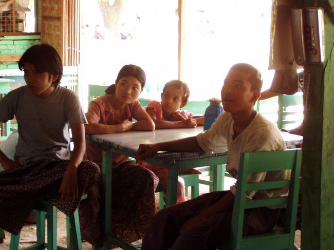 　兄弟揃って真剣な表情でテレビを見ている。奥左から、下から２番目の妹。中学浪人(２浪中)。この調子じゃ今年<br />もだめだな。<br />　右、ウィンウィンの下の妹。石のように無表情。中学で勉強は投げた。たまに幼い子を泣かしている。<br />　一番右、レッパンチバウ村から来た女の子。<br />　手前。末弟のミンミン(愛称)。口を開けて間抜け面だが、実はキムタク系のいい男。学校が休みなので帰省中。<br />　それにしてもそんなに面白いかあ？「チャングムの誓い」。<br />　アウンヤダナーレストランは、ウィンウィンが実質的な店長で妹のエティダが副店長みたいな感じ。８人兄弟姉妹の中でこの二人が一番しっかりしている。ウィンウィンは３番目で、一番上のお兄さんもレストランと店の前にある電話屋を手伝っている。公務員だったとか、ホテルで働いていたが居眠りしていたのを見つかってクビになったとか、いろいろな噂がある。ウィンウィンによると酔っ払い対策だそうだ。バスステーションが近いし、馬車の御者もしょっちゅう食べに来るから、当然酔っ払いも出るのだろう。そんな奴らを女性だけで相手をするのは確かに大変だと思う。<br />　次女は結婚してヤンゴンに住んでいたが、だんなさんが新首都ピンマナーへ単身赴任となったので、幼い娘を連れて帰ってきており、彼女も店を手伝っていた。食い物がいいのか、兄弟姉妹の中では一人だけ丸々と太っていた。<br />　六女(？)は、この時司法試験を受けるためにヤンゴンへ行っていた。タラバーゲートホテルで働いていたが、試験のためやめざるを得なくなったという。個人的にはこの姉妹の中ではキュートで一番かわいい...と思う。今回会えなくて残念だった。<br />　結局、７人の兄弟姉妹がここで働いていた。ほとんどの時間、客の数より店員の数の方が多く、日本でこんな店があったら、１ヶ月でつぶれてしまうだろう。もっとも、家は狭いし、レストランにはテレビもあるし、こっちの方が快適なんだろうが。