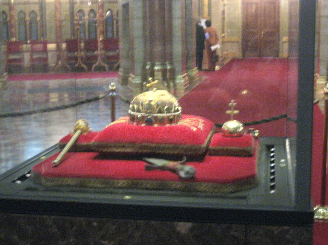 ハンガリーの聖なる王冠<br /><br />最大ズームにしてこの大きさです。<br />ちょぃとブレているのはお愛嬌。<br /><br />私がお腹に抱えているタウンバックが大きいので、写真を撮るためにちょいと乗り出したら、荷物だけ進入禁止のロープを越えてしまって、注意されてしまいましたよ@<br /><br />この王冠のレプリカが王宮のマーチャーシュ教会にあるので、近くでじっくり眺めるのはそちらを楽しみにするとしましょう。<br /><br />レプリカの写真<br />http://4travel.jp/traveler/traveler-mami/pict/11491766/<br />関連の旅行記<br />http://4travel.jp/traveler/traveler-mami/album/10104710/<br /><br />国立博物館で買った冊子「ハンガリー歴史アルバム」によると、聖イシュトヴァーンがキリスト教導入を決定する前に、ローマ・カトリック教皇とビザンチン皇帝の両方から王冠を贈られたそうです。<br />この「ハンガリーの聖なる王冠」は、最終的にはローマ教皇より贈られたものと伝えられています。<br /><br />この王冠は1918年、ハンガリー王国解体まで使用されました。<br />第二次世界大戦後、国外に持ち去られ、長い間アメリカに保管されていましたが、1978年にカーター大統領の決定によりハンガリーに返還されました。<br />当初は国立博物館にあったそうですが、2000年からこの国会議事堂に移転されたそうです。<br /><br />てっぺんの十字架が曲がっているのは、２年前のバスツアーガイドは、昔、王冠が騒動で行方不明になって再び見つかったときにはひん曲がってしまっていたが、これを元に戻すのに失敗したりしたら、と誰もが恐れ多く修復したがらなかったので、そのままになってしまった、などと説明してくれました。<br />今回は、戦時中、ブダペストが攻撃されたとき、これを安全なところに避難させようとした者が用意した入れ物が少し小さくて、無理していれたら曲がってしまったせいだ、とか、イシュトヴァーン王はキリスト教の導入を決めたとはいえ、キリスト教や西欧のことに詳しいわけではないので、十字架とはこのように曲がっているものだと思い込んでそのまま使ったのだ、などと冗談だか本気だかわからない説明を聞きました（議事堂のガイド以外からも）。<br />諸説あるということでしょう。
