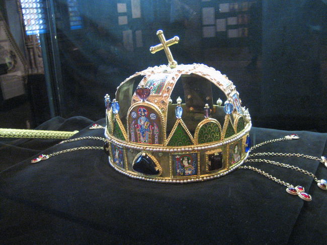 マーチャーシュ教会の宝物館にあったハンガリーの聖なる王冠のレプリカ<br /><br />これももう一度じっくり見たいと思っていました。<br />２年前は、なぁーんだ、レプリカじゃん！<br />と思ったのですが、すごく良くできているんです。<br />それに、国会議事堂の本物は、近くでじっくり眺めることはできませんでしたから。<br /><br />このレプリカを作るにあたって、聖なる王冠の研究結果や解説のようなものが部屋に展示されていました。<br />２年前はあまり興味がなかったのですが、今回は興味が沸いたため、展示をじっくり読むよりその内容が書かれていると思われる2003年刊行の “The Holy Crown of Hungary”という冊子をマーチャーシュ教会のギフトショップで買いました。<br />値段は1,000フォーリントでした。<br />（2006年10月現在、1フォーリント＝約0.6円）<br /><br />この冊子は写真もとてもきれいです。内容も後でざっと読みましたが、なかなか興味深かったです。<br />以下はその内容からの抄訳のようなものです。<br /><br />聖なる王冠は、ハンガリーの昔の法典では、王その人と王冠が示す王権とは別の存在で、王はあくまで聖なる王冠を通じて王権と統治権を授かるということになっていたそうです。<br />その顕著な例として、王冠の名の下に投獄された王もいるくらいです（ルクセンブルク家のジグムント王）。<br /><br />聖なる王冠についての一番古い伝説は、1000年にローマ教皇から初代国王イシュトヴァーンに贈られ、イシュトヴァーン王のエステルゴムの戴冠式のときにかぶられたというものです。<br /><br />ただし、この王冠は、エナメル象眼でビザンチン皇帝の肖像が描かれた下のギリシャ風の王冠部分と、上の十字に交差し、斜めになった十字架が乗っているラテン風の王冠部分とが合体してできたもので、上の部分だけがイシュトヴァーン王と関わりがある部分ではないかとの研究結果もあるようです。<br /><br />聖なる王冠についての研究が最初に始められたのは、1790年にハプスブルグ家のレオポルド２世がウィーンに持ち帰ったときからだそうです。<br />一番最近の研究は、1978年にアメリカからハンガリーに返還されてからです。<br /><br />聖なる王冠が作られた時期については、諸説あって、5世紀から15世紀までと幅広<br />く、ずっと信じられてきた伝説、すなわち1000年にローマ教皇から贈られたという説を完全に否定する材料はないようです。<br /><br />象眼で描かれている像は、下のギリシャ王冠には、全能の支配者たるイエス・キリスト、受胎告知の天使ガブリエル、最後の審判の天使ミカエル、聖ジョージ、聖デメトル、聖コズマ、聖ダミアン。<br />それから世俗の王が３名：キリスト教を信仰したビザンチン皇帝ミカエル・デュカス（Michael Ducas）とその息子のコンスタニイネ（Constanine）。同じくキリスト教を信仰したトルコのゲオヴィカス（Geobicas）。このトルコの王は、昔西欧ではハンガリー人をトルコ人といっしょくたにしていたので、イシュトヴァーン王の父ゲーザ公のことではないかという説もあるようです。ギリシャ文字でそれぞれ名前も書かれています。<br />ただしこの世俗の３名の王の部分は後から入れ直した細工の後があるので、デュカス王の部分は、当初は、イエス・キリストと対をなすようにマリア像が描かれていたのではないかといわれています。<br /><br />上のラテン王冠部分には、キリスト、それから教会の礎であり印の鍵を手にしている使徒ペテロ、福音書書記者でもあるヨハネ、宣教師パウロ、大ヤコブ、アンドリュー、フィリップ、トマ、バーソミュローですが、バーソミュローはかなり昔に破損してしまったため、レプリカでも復元されていません。それがバーソミュローだと分かったのは、それぞれみなラテン文字で名前が書かれていたからです。<br /><br />写真では、ちょっと見づらいですが、下のビザンチン風の部分の「全能の支配者たるイエス・キリスト」のエナメル象眼の絵を中心に写しました。<br />私のように冊子を買うまでもなく、壁の天使や聖人の絵の拡大写真をぜひご覧になってください。<br />なかなか可愛いですよ@