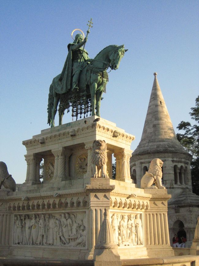 【3631】<br />夕方のやわらかな陽を浴びた、三位一体広場の聖イシュトヴァーンの騎馬像<br /><br />広場の騎馬像なんてお約束な写真を撮っても……と思うのですが、撮りたくなるものですね。<br />記憶にあったのよりも、ずっとかっこいいと思いました。<br />その格好良さと迫力がどこまで写真に収められるか。<br />背景に漁夫の砦のとんがり頭の１つを入れてみました。<br /><br />騎馬像のイシュトヴァーンはアールパード家の紋章に使われていたダブル・クロスを掲げています。<br /><br />おまけ<br />2004年に撮った三位一体広場の写真<br />http://img.4travel.jp/img/tcs/t/album/src/10/03/88/src_10038899.jpg?00000000000000<br />関連の旅行記<br />http://4travel.jp/traveler/traveler-mami/album/10031269/<br />王宮地下迷宮の迷冒険記！あり。