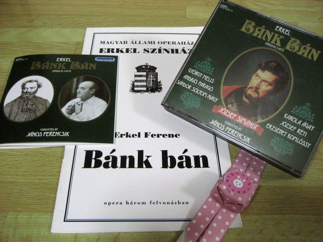 「バーンク・バーン」のプログラムとCD（ケースと中の解説書）<br /><br />プログラムは340フォーリントとそれほど高くなく、英語であらすじと出演者リストが載っているので、買ってしまいました。<br />（2006年10月現在、1フォーリント＝約0.6円）<br /><br />といっても、プログラムに英語が併記されているのはあらすじだけでした。作品や作曲家の解説も長々とあるようですが、そちらはハンガリー語オンリーでした。<br />とはいえ、すべて英訳がついていても、私に読み切れたかとどうか。<br /><br />プログラムの出演者リストは、重要な役柄順ではなく、身分の高い順番から書かれているのは面白いと思いました（少なくとも日本では重要な役柄順というのがふつうだと思います）。<br />なので、最後にちょこっとしか登場しない王様アンドラーシュ２世がまずトップに挙げられていて、いわば悪役である王妃やその弟オットーが、主役のバーンク・バーンより先に並べられています。<br /><br />CDは後日、オペラ座ギフトショップに舞い戻って買いました。<br />公演を見て、「バーンク・バーン」の音楽がとても気に入ったので、帰国したらAmazonかCDショップで注文しようと思ったのです。<br />しかし、考えてみたら、「バーンク・バーン」はハンガリーの国民的なオペラなのです。日本で注文するより、ハンガリーで買った方が早いのではないでしょうか。<br />というわけで、その後、ブダペスト市内にある大型ショッピングセンターのCDショップを回りましたが、エルケル・フェレンツのCDはあれど、「バーンク・バーン」は見つかりません。むむむ。<br />結局、オペラ座に舞い戻り、併設のギフトショップで買いました。<br />価格は定価の7,200フォーリントでした。<br />CDショップなどで見つけることができたら割引価格も夢ではなかったかもしれません。<br />実際、西駅に隣接している「ウェストエンドシティセンター」という大型ショッピングセンターのミュージックショップで、代わりにオペラ「聖イシュトヴァーン」なるCD２枚組を見つけたので買ってしまったのですが、値札には8,390フォーリントとあったのに、レジに通したら1,990フォーリントでした。<br /><br />プロローグで紹介したサイトにも「バーンク・バーン」のあらすじがありますが、ここでは私が買ったプログラムからも抄訳でざっくりとあらすじを紹介しましょう。<br /><br />第１幕<br />国王アンドラーシュ２世が遠征で留守中の宮廷。外国出身の王妃ゲルトルーディスが女主人役を務めていましたが、彼女は外国人を優遇し、また浪費を尽くしていました。副王の地位にあるバーンクは、国王に代わって国内の貧窮ぶりを視察していましたが、その留守に、王妃の弟のオットーが、バーンクの妻メリンダをモノにせんと狙っていました。外国人の王妃とその弟がのさばる宮廷に不満を抱き、国を憂えるハンガリー人貴族のリーダー、バーン・ペトゥールは、反乱計画を立てており、地方の視察から戻ってきたばかりのバーンクに参加を呼びかけます。オットーがメリンダを狙っていることも告げ、このまま王妃の専横をのさばらせて良いのか、とバーンクを説得しようとします。バーンクはあくまで王権に忠実であろうとします。しかし、オットーが実際にメリンダに言い寄るところを目撃して（ひいては国の重鎮であるバーンクに対する無礼ということになるでしょうか。オットーの言動はハンガリー国内での外国人の横暴の象徴のようです）、ペトゥールとその仲間たちの会合に出席することにします。<br /><br />第２幕第１場<br />ヴィシェグラート城の入口で、年老いた農民ティボルツが、外国人に蹂躙された国の窮状をバーンクに直訴します。ところがバーンクは妻やペトゥールのことを含めてこれから自分がどうすべきかで頭がいっぱいだったので、話半分にしか耳を貸しませんでした。そのころ、オットーは姉の王妃ゲルトルーディスの容認のもと、ついにメリンダに実力行使します（未遂ではないかと思ったのですが、よく分かりませんでした）。傷ついたメリンダは半狂乱になってバーンクに懺悔します。バーンクは幼い息子に八つ当たりしてしまいますが、すぐに我に返ります。結局、バーンクはティボルツに付き添いを頼み、息子と妻をハンガリー東部の自分の領土に帰すことにします。<br /><br />第２幕第２場<br />宮廷で、バーンクは王妃ゲルトルーディスに国の窮状と妻に対するオットーの無体な行為の責任を問いつめます。王妃はバーンクをバカにして自分の所業を認めず、短剣を取り出します。２人はもみ合っているうちに、バーンクは王妃を刺し殺してしまいます。<br /><br />第３幕第１場<br />ティボルツはバーンクの領土に戻るべく、メリンダと彼女の息子を連れてティサ川を渡ろうとしていました。ところが精神がすっかり病んでしまったメリンダは、ティボルツが目を離した隙に、息子と共に川に身を投げてしまいます。<br /><br />第３幕第２場<br />宮廷では、王妃の棺の前で、国王アンドラーシュ２世は宮廷貴族たちにその責任を追及しました。貴族たちが無関与を主張する中で、バーンクは、王妃の非道ゆえに彼女を暗殺したと白状します。国王とバーンクが今にも剣を交えようとしていたところへ、ティボルツがメリンダと息子の遺体と共にやってきます。バーンクは剣を落とし、妻子の遺体に嘆き伏しました。<br /><br />おわり<br /><br />あらすじだけ追うと、バーンクがもう少し妻のことを気にかけてあげれば、最後の悲劇は防げたのではないかと思わなくもないのですが（字幕がないので確信できませんが、見ていた限りではオットーに乱暴されたメリンダをバーンクが責めるようなシーンがなきにしもあらず）、義人バーンクの頭の大半を占めていたのは、あくまでハンガリーという国の将来だったのでしょう。<br />まあ、個人の尊厳や女性の地位よりは国の運命とハンガリー人貴族としての名誉の方が優先されるのが当然な時代だったでしょうから、仕方がありませんけれどね。