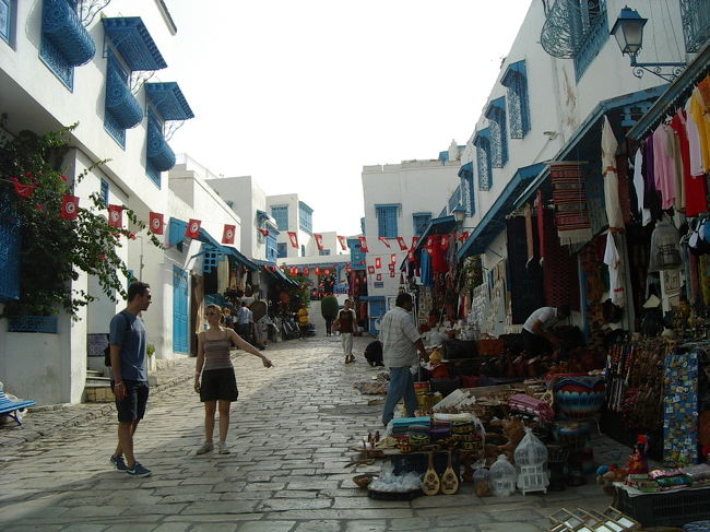 土産物屋が並ぶハディ・ザブルーク通り。白とチュニジアンブルーの建物に、赤い国旗がなびきます。<br />