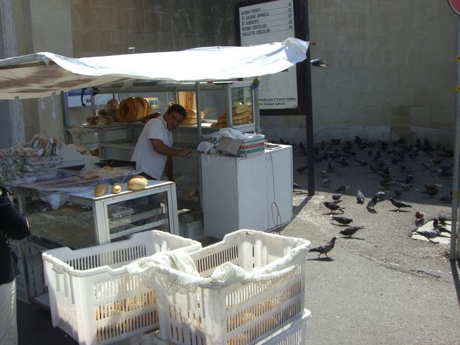 ヴァレッタのバスターミナル付近の露天のパン屋さん。確かにマルタのパンは美味しかった。出勤途上の勤め人たちも買っていく。屋台の裏にはハトの大群。店のオヤジがパンくずをまき与えるのが習慣の様子。マルタの人は生き物に対して優しい心根を持っているように感じる。<br /><br />ヴァレッタはそもそもが要塞であって、それほど樹木は多くもありません。だからこそなのか、家々の玄関やベランダには鉢植えが置かれていて、丁度東京の下町の路地にも似た風情があります。そんなヴァレッタの石畳の階段を歩いていたところ、食器洗剤の容器のようなものを手にしたオヤジが家から出てきた。何気なく様子を見ていると、石畳の間から顔を出している野草（雑草）にピュッ、ピュッと水をまいて引っ込んでいった。まるで毎日の日課かのようにそそくさと。邪魔ならヒョイと抜ける小さな草ゆえに、決して除草剤などではなく、間違いなく水を草に与えたのだと思う。道行く人にいつ踏みにじられても不思議ではない紛れもない雑草。マルタの人の心根の優しさを見た思いがした何気ない瞬間でした。<br />