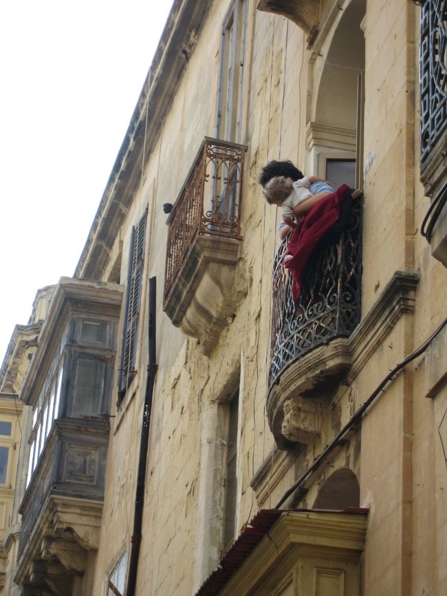 出窓から景色を眺めるのがこの町に住む人々の朝の慣わしのよう。顔見知りと「オハヨー！」と声を掛け合う。そんな上階の人たちから、「ちょっとあんた、後ろからクルマが来るよ、気をつけなよ」と何度となく声をかけられました。いや、言葉は分からないのだけれど、きっとそう言ってくれているに違いない。下町の人情。世界遺産だけど、決して死んではいない街。生身の人情が通った生きている世界遺産の街・ヴァレッタ。<br /><br />もともと狭い道に、路肩にまで乗り上げた状態で両サイドに路上駐車のクルマが並ぶ。この街の人は世界一縦列駐車が得意な人たちに違いない。歩道も半ば奪われている歩行者は少しばかり注意が必要。