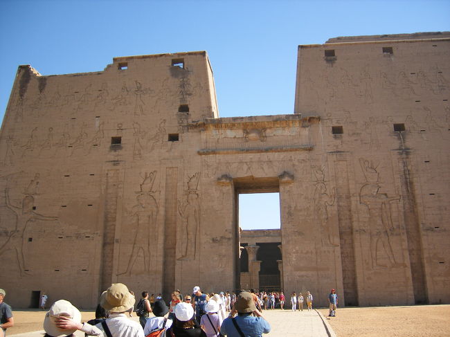 ホルス神殿・塔門<br />プトレマイオス４世の時代、至聖所と第１列柱室の一部など神殿の北区域が完成。その４０年後プトレマイオス８世の時、建築が盛んになり前２３７年８月２３日、神殿竣工式が行われたとの碑文がある。しかし実際にはその後１９５年かかって完成した。<br />その塔門は、幅１３７Ｍ、高さ３６Ｍ、ルクソールのアメン神殿第１塔門に次ぐ大きさである。壁面のレリーフはプトレマイオス１２世がホルス神とハトホルス神の前で敵をうちすえている。