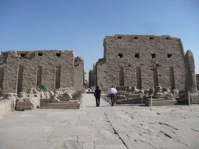 ♪♪♪====カルナック神殿/Karnak Temple=====♪♪♪<br /><br />カルナック神殿は中王朝時代にテーベの守護神のアムンとラーが合体してアムン・ラー神としてあがめられるようになった第13王朝時代に建築が開始された。テーベの神殿としてはこちらが本殿となり、後代々の王が寄進、増築を進めたため、東西に走る身廊部分はルクソール神殿と同じ様式なるも、そこから外に広がるようにいびつな形で広がっていっている。<br /><br />●羊頭のスフィンクス参道と第一塔門<br />カルナック神殿の身廊は東西へと延びているが、その西側第1塔門の前に延びるスフィンクスの参道。長さは52メートルにも及ぶ。