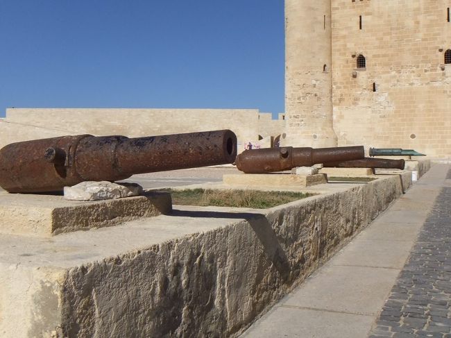正面からの全体写真を撮り忘れてしまいました。(動画や記念写真には写っているのですが、ここでは公開しません。)この要塞は、ファロスの灯台の跡地に建てられた特別なものです。ファロスの灯台はアレクサンドロス大王の案でプトレマイオス２世により紀元前２８０年頃に建てられたとても高い灯台で、当時の世界七不思議の１つでした（今残っている世界七不思議はピラミッドだけ）。高さ１２０ｍで、５０km以上先からも見えたそうです。灯台は、１層目が正方形、２層目が八角形、３層目が円柱形というデザインだったそうですが、１３７５年の地震で崩壊してしまったそうです。その後、灯台の石材を利用して、１４７７年にマムルーク朝のスルタンだったカイトベイにより、十字軍の攻撃を防ぐ要塞が建てられたのだそうです。カイトベイでのガイドさんの説明を聞き終わった時点で１１時２０分。１１時４０分までの２０分が自由時間です。要塞ですから、当然大砲があります。要塞前の道の左側に並んでいます。