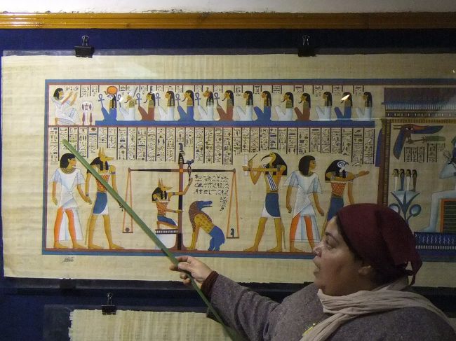 店内には綺麗な絵の描かれたパピルスが並んでいます。主なものをガイドさんが説明。絵はちゃんと、古代エジプト時代に描かれていたデザインです。この絵は死後の世界を表しています。一番左のファラオ（王様）は死後、アヌビス神（ジャッカルの神様）につれられて、裁判を受けます。ここには天秤があって、右側には聖なる羽（マアトの羽といいます）がのっています。左側にはファラオの心臓をのせます。この時、この絵のようにファラオの心臓の方が軽ければ、生前よい行いをしていたということで、天国に行けます。しかし、もし心臓の方が重かったら、悪いやつだとみなされ、中心棒の右側のワニとライオンが合体したような怪獣（アメミットという名前です）に食べられてしまいます。裁判の様子は、天秤の右側のトト神（トキの神様）が記録します。