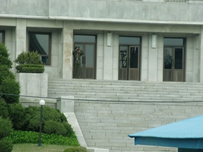 北の板門閣に、軍人さん1名確認できました。<br />双眼鏡で南側を監視しています。<br /><br />確認できたのは1名だけでしたが、南北それぞれ35名が常駐する決まりになっているそうで、建物内から南に向かって銃口が向けられていることも否定できないとのこと。<br />だから、北に向かって手を振ったり、指さしたりすることは厳禁です。意味ありげなことをすると、「あそこを標的にせよ」と指示が出たと判断され、即座に反撃があってもおかしくない。それほど緊張した場所でした。