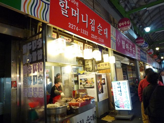 『ハルモニチッスンデ』<br />住所：鍾路区鍾路5街広蔵市場 東門18号 <br />http://www.seoulnavi.com/food/2649/<br />広蔵市場の斜めの十字路から少し東寄りにあるスンデ専門店。<br />酒盛り中のおじさん達で混んでるけど、女1人だけど、ひるむなノラ！<br />いざ突撃〜！<br /><br /><br />