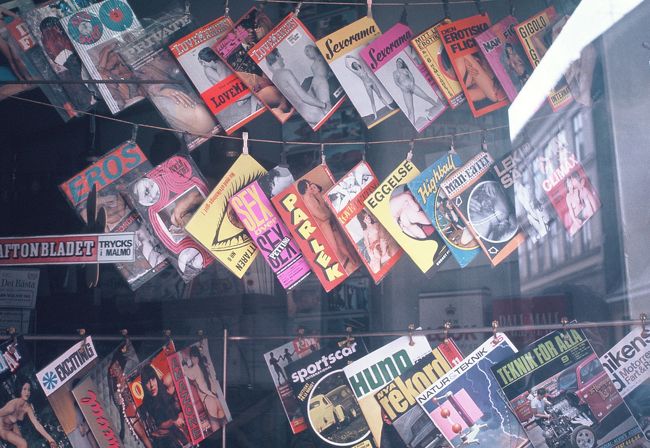 ポルノ雑誌(pornography)の店。<br /><br />７０年代にはアメリカも開放的になりましたが、６０年代にはポルノ雑誌がスタンドで売られるようなことは未だ無かった。<br /><br />６０年代、セックスに関しては最も開放的だったのはヨーロッパ、特に北欧でした。　　　このような雑誌をアメリカに持ち帰ったら、税関で没収されたと思います。<br /><br /><br />さて、日本語、ポルノ、の語源になっている英語は　pornography。　　英語の語源はギリシャ語の娼婦 porne。　　その語尾に 書く　を意味する　graphy　を付けた言葉。　　<br /><br />「そうか、英語で娼婦は　ポルノ　なんだ」<br /><br />いや、娼婦は　英語で　prostitute です。　　ラテン語の語源で　pro が前、statuere　が立つ。　　前に立つ（者）が娼婦なのですね。<br /><br />脱線します。　　英語で賛否　は　pro　と con　ですね。　　そこから生まれた冗談が、　若し、prostitution が　売春だとすると、その反対になる constitution　は 何か？<br /><br />勿論、アメリカの憲法です。<br /><br />面白いのは英語で　"take a constitutional"　と云うと　散歩をする　と云う意味になることです。　<br /><br />