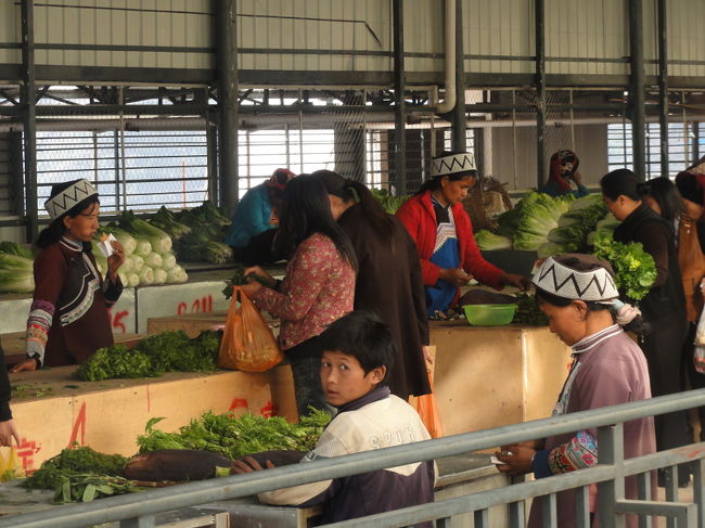 　緑春の中で最も民族衣装を見かける場所が、町の中心にある市場。道路を挟んで老百姓農貿市場と緑春商業広場の２つのマーケットが向かい合って建っています。注目は緑春商業広場奥の建物二階にある野菜売り場。自ら育てた野菜を売るオバサン達の民族服着用率はなんと8割以上。今までの市場めぐりは何だったんだ!という感じです。<br /><br />写真: 野菜売り場の端のグループだけ帽子が違う。<br />