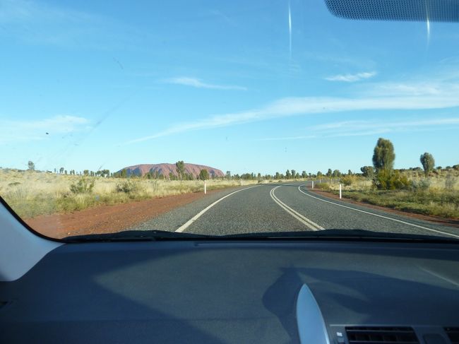 買い物を終えたら一旦部屋へ荷物を置きにいき、ウルル・サンセット観賞をしようといざ出発。<br /><br />エアーズ・ロック・リゾートを出て南へ車を走らせます。私が運転し、助手席の嫁が写真を撮りました。目指すはサンセット・ビューイング・エリア。大きな一枚岩が段々近くなってきます。