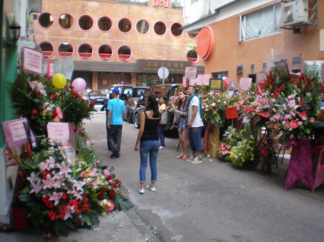 せまい道の両側に数多くの花輪（日本のとはちょっと違う）を並べて開店祝いのイベントをやっているところに遭遇。｢プリティリトル｣というかわいい名前の若い人向けのファッションの店でした。