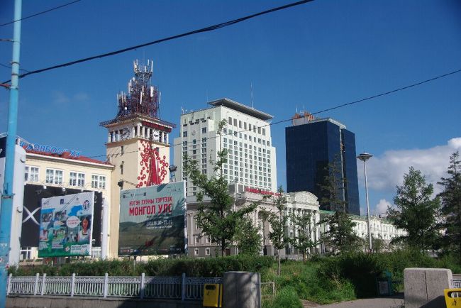 　朝９時，私たち一行を乗せた２台のワゴン車は，一路３００キロ先のブルドを目指して出発しました。<br />　車窓の風景その１（スフバートル広場前）です。公園の樹木の向こうには，昨夜不思議なライトアップで迎えてくれた市庁舎が見えます。