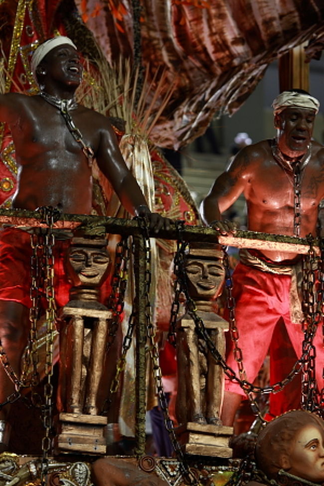 サンバのチーム、エスコーラは、リオのスラム　ファヴェーラにて誕生しました。1888年の奴隷解放前、リオの見栄っ張りのプランテーションの主人は奴隷を着飾らせて御者や従者にしたそうで、奴隷解放後、田舎の解放奴隷はリオに行けばああいう暮らしができるのではと期待して、リオに押し寄せたそうです。そういう解放奴隷達が集団で暮らしたのがファヴェーラ。1940年代、1970年代と経済危機が起こるたびに、黒人のみならず白人貧困層もここに住み着きファヴェーラは拡大。ブラジル最大のファヴェーラはリオのイパネマ海岸の南端から丘の上に広がる地域だが、リオではあちこちの丘の中腹に、お屋敷街が突然、積み重なる小さなブロックのようなカラフルな家々の連なり、ファヴェーラに変わるのが見受けられます。