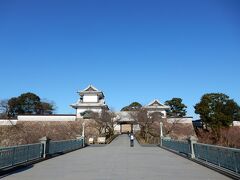 金沢駅改札を出て、バス停を探していると、もてなしドームの左側にあるのにすぐに気づいた。停まっているバスに乗車、すぐに発車だ。ナイスタイミング！　兼六下バス停まで１５分程度だったか？
バス停から兼六下交差点に戻ると、右側に金沢城の石垣と石川門が見える！　おお憧れの金沢城だ。左側は兼六園。兼六園への坂道を上がりつつ、心は金沢城へ！　兼六園入り口から見る金沢城石川門（現存）。快晴の空をバックの石川門、素敵だ！
駆け寄りたい気分を抑えて、予定通り、ますは兼六園だ！　観光客にお願いしてふたりの記念撮影していただいた。