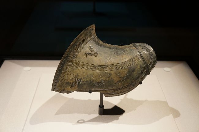 獣紋飾板（初期鉄器～原三国時代・前3～前1世紀）伝韓国慶州出土<br />《東京国立博物館・解説より》<br />周縁に鋸歯紋と格子紋を配し，その中に双頭渦紋と鹿，犬が陰刻される。鹿の一頭には棒のようなものが刺さっているが，先端が二股に分かれて矢羽根のようである。狩猟の場面であろう。朝鮮の鋳銅技術の高さを示す作例であり，いわゆる異形青銅器の一種で，祭具として用いられたのだろう。皮袋の飾り，または，肩を覆う鎧の一種とも考えられている。 