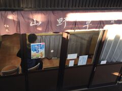 松本駅構内の跨線橋から0番線ホームに向かって降りると、階段の影に1軒の立ち食い蕎麦屋がある。
店の名は「山野草」。
店内は程よい狭さで、どこにでもある駅蕎麦のよう。
だが、その正体は知る人ぞ知る名店。
ここは通常の立ち食い蕎麦と、特上の２種類ある。
特上は生麺から茹でるため、予め茹でてある通常バージョンより時間がかかり、しかも高い。
それでも客の注文を見ていると、ほとんどが特上を注文している様子。
もりそばでも40円程度の差額しかないので、たとえ待たされても特上のほうがお得感はある。
ここで「特上きのこ山菜そば」を注文。
蕎麦粉もキノコも山菜も地元信州産かどうか分からないが、そこは気分というもの。
それに蕎麦そのものも普遍的な立ち食い蕎麦より明らかに美味い。
手頃な価格で信州気分を満喫できたのだから野暮は言いっこなし。
旅の合間に心の隙間を満たされた思いで跨線橋の階段を登った。