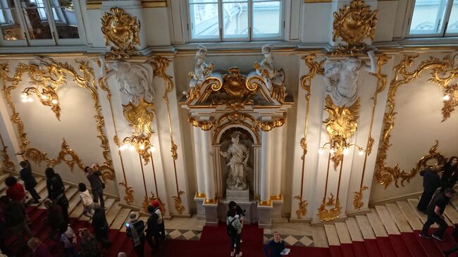 10:49　やっぱり混んでるわ、すでに人だかり。頭上のティツアーノだけは頑見しとこ。金箔の貼り方がペテルゴフと比べものにならない丁寧だ。ここら辺にインフォあって無料の館内図あるんではないのか。見当たらないんだけど<br /><br />冬宮中央階段《ヨルダン階段》<br />冬宮の中央階段はB.=F.ラストレッリの設計によって建設された。1837年の火災後、この復元に当たったV.=P.スターソフはバロックの内装をそのまま残した。最初の踊り場までの部分は厳かにほの明るく、その後に続くその後に続く階段主要部とは対照を成す。そこでは光があふれんばかりの空間が鏡の中に反射を繰り返しながら横へと、そして天井画の偽りの空に向かって上へと、増幅するかのように広がっていく。18世紀、階段正門には金箔の塑像が飾られ、手摺子にも金箔が施されていた。そして、2階の柱廊天井は、組になったバラ色の人工大理石からなる円柱に支えられていた。内装の復元に際してスターソフは何か所か変更を加えたが、特に光量の対照を強める目的で下段の窓を作った。バラ色の円柱に代えて、一枚岩から掘り出された鼠色のセルドボリ産花崗岩の円柱を設置し、木彫の手摺子の代わりに重厚な石手摺を配した。燃えてしまった天井画の代用品として、スターソフは冬宮の倉庫にしまわれていた中から、一時冬宮の一室を飾っていたという18世紀のイタリア画家ディッツィアーニによるオリンパスに集うギリシアの神々を描いた絵を選んだ。（土産本）<br />