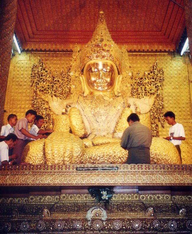 マンダレーの市内に入る。<br />マンダレーはビルマ族コンバウン王朝があった古都。ミャンマーのほぼ中央に位置し、ヤンゴンに次ぐ第2の都市だ。大英帝国はインドを拠点にアジアの植民地化を進めていたが、隆盛期のミンドン王が逝去した後、1885年にティーボー王を捕らえてマンダレーを手中にした。王都としてはわずか25年で幕を閉じた。<br />　<br />「マハニムバゴダ」には、有名なマハニム仏が安置されていた。<br />この仏像は、マンダレーの中で最も大きく高さ4mもある。創建は1784年で、パガンの寺院群と比べるとそんなに古いものではない。参拝者が金箔をこの仏に貼り、願をかけている。そのため随分着ぶくれ状態が進んでいる。仏が安置されている内陣は女人禁制になっており、仏に触れることはできない。<br />この境内には、アンコールワットから略奪されたブロンズの像が安置されていた。その経過をたどると、当時の勢力争いの一端がうかがえる。1400年代にタイの軍勢が当地から略奪をして後、モン族がこれを略取しバゴーに移す。その後も転々とし、このバゴダに納められたのだ。<br /><br />