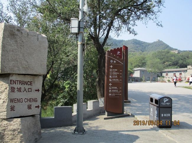 ↑ 歩行登城入口の前の広場。（望京文化広場前）<br />　写真の右手の低い稜線に長城が目に入る。<br />　バス降車場から検査場をとおり、約700mを歩いてきたところ。<br />　バスを降りたすぐ前に切符売場（滑車用と思う。）があったが、それを左手にやり過ごし、スマホにダウンロードしていた地図をたよりに、バス降車場の裏手へと続くなだらかな登り道（景区公路）をゆっくり１０分ほどすすんで検査場到着。<br />　検査場ではライターこそ上納したものの、ここから先の登城口では、当方は６０歳以上なので入場料金は納めずに済んだ。<br />　これより先、幾度も老人優待を受ける。<br />　（日本ではこれほどの優待はなかったのでは？）