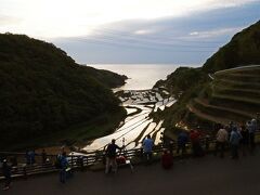 眼下に広がるのは日本の棚田百選選定された、夕日の名所として知られる玄海町の“浜野浦の棚田”。
玄界灘に面した棚田は、毎年4月中旬から田んぼの水張りが始まり、5月上旬には田植えが終わるため、GWは棚田夕日鑑賞にピッタリのタイミングだったので、是非訪れたかった場所…天候不良も考慮して唐津での滞在を2日間確保したのはこのためだった。
展望デッキがある広い展望台には、手摺に沿って三脚を構えて陣取るカメラマンが占めていたけど、コロナ禍じゃなければもっと混んでいたかも。
この時間でも太陽はまだ高い位置にあり、地平線周辺を分厚い雲が覆っているので、下段の展望デッキまで降りて様子を見ることにした。