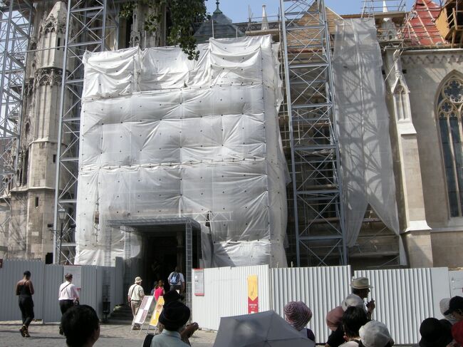 シートに覆われた出入口から入場。<br /><br />マーチャーシュ教会の正式名は、聖母マリア教会。<br />元々は1015年に、国王イシュトバーン1世が建造した教会が起源で、<br />13世紀にモンゴルに破壊され、その後ベーラ4世の命により再建。<br /><br />14世紀後半から15世紀に、<br />マーチャーシュ王によってゴシック様式に大規模改築が行われてからは、<br />マーチャーシュ教会と呼ばれるようになった。<br /><br />16世紀にはブダがオスマントルコに占領され、内部がモスク様式に塗り替えられた。<br />そして、19世紀末の二重帝国時代になって大掛かりな増改築をし<br />カトリック様式に戻され、現在の姿になった。