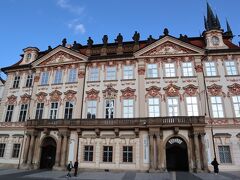 「石の鐘の家」の隣は、「ゴルツ・キンスキー宮殿」です。
「ゴルツ・キンスキー宮殿」は、もともとは別の3つの建物でしたが、1508年に当時の所有者であった政治家「アルブレヒト・フォン・コロヴラート」によって1つの建物に統合され、1560年には別のオーナーによってルネサンス様式の建物として拡張工事が行われました。そして1755年から1765年にかけて、「ゴルツ伯爵」によって現在の建物の外観に改装され、1768年に「ゴルツ伯爵」が亡くなると、オーストリア生まれの資産家「フランツ・ウルリヒ・キンスキー」がこの建物の所有者となりました。
現在の建物名は、「ゴルツ」と「キンスキー」2人の所有者の名前に由来して命名されたものです。

ロココ調のスタッコ細工で飾られた窓やペディメントをはじめ、屋上にそびえる彫像など、旧市街広場に立つ建物の中で、一番華やかな建物です。
