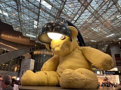 まずセンターのアライバルロビーに行き、そこから伸びるCコンコースに向かいます。ハマド国際空港は、2015年頃に来たことがあります。当時はまだ何もない空港でしたけれど、ワールドカップもあってずいぶん整備が進んだようで、雰囲気がずいぶんと変っていました。アイコニックな黄色い熊のオブジェは、なんでもニューヨークのアートなんだとか。

「こんなものあったっけ？」
「記憶ないねえ？前来た時はまだ、ゲートの南北繋ぐ電車が工事中で乗れなかったり、工事中ばっかりだったもの」
「この熊、有名みたいだよ。ほら記念撮影してる人がたくさん」
「ふーん」