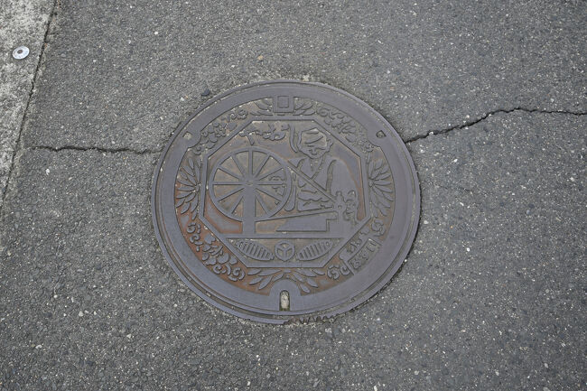 八尾市の汚水桝の蓋。<br /><br />「河内木綿」伝承の地である八尾市を象徴して、八角形の内側に糸車で糸を紡ぐ姉さんかぶりの女性を描き、その周囲に市の花「キク」と木綿の実がデザインされています。