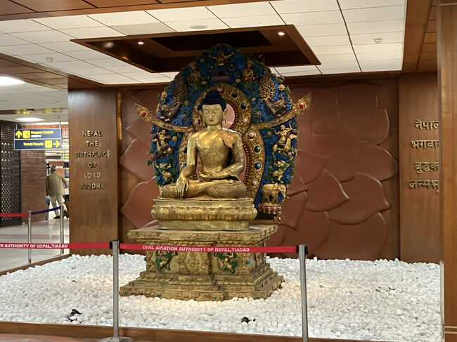トリブバン空港へは、空港混雑のためか上空で何回か旋回したりして、予定（16：30）よりもかなり遅れて、18時前に到着。ボーディングブリッジの無い空港なので、バスでターミナルに移動して仏像がお出迎えです。<br />日本との時差は３時間15分。うん？　何だよ、この15分というハンパな数字は!!<br />公式見解では、ヒマラヤ山脈のガウリ・シャンカルという山にちょうど子午線がかかるのでそれを避けたためだそうですが、インド（インドとの時差は３時間30分）との差別化を図るためわざわざ15分の差をつけたらしいですね。いろんなオモワクがあるもんだ。